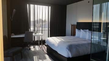 A hotel room with a large bed, a desk with a chair, a small table with a lamp, and a view through the window.