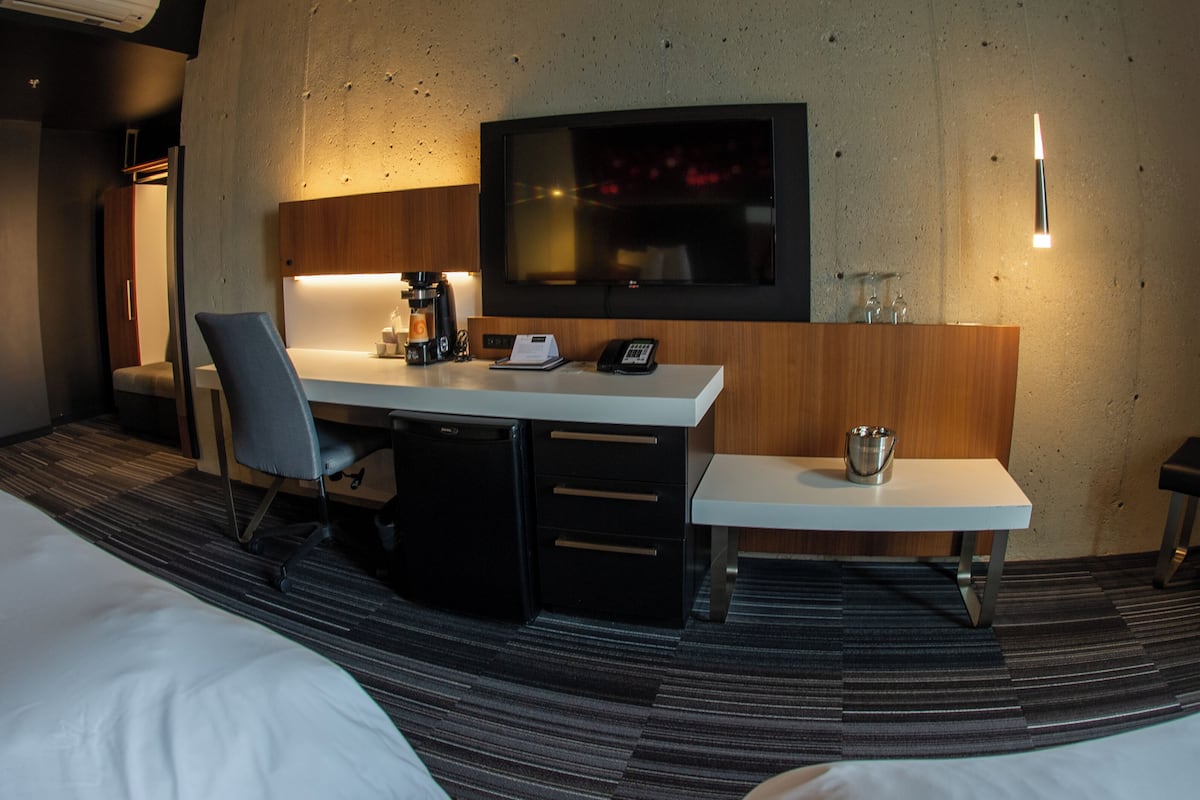 A hotel room with a bed, a desk with a chair, a television, a coffee machine, and a small table with a bucket.