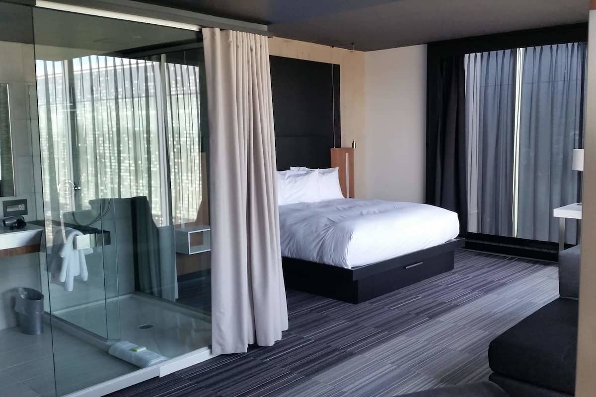 A modern hotel room with a large bed, a glass shower enclosure, a bedside table, and a view of the city through the windows.