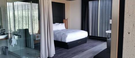 A modern hotel room with a large bed, a glass shower enclosure, a bedside table, and a view of the city through the windows.