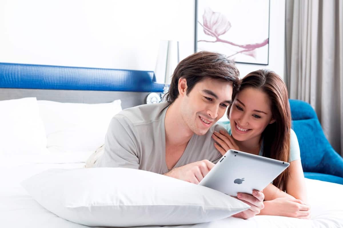 Una pareja recostada en una cama con una tableta, un cabecero azul y un cuadro en la pared.