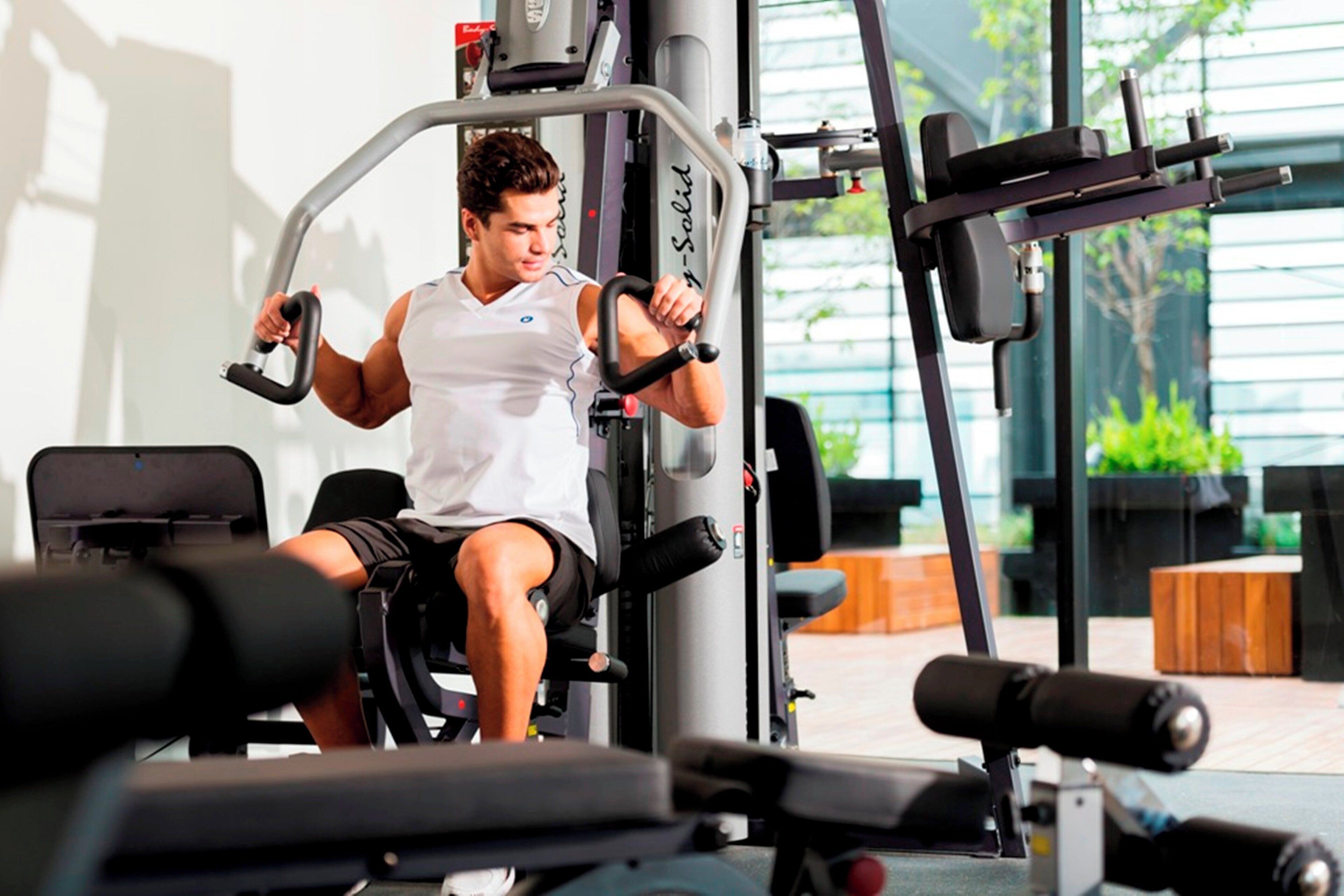 A person using a gym machine with a weight stack and handles, in a room with exercise equipment and a window with greenery outside.
