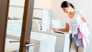 Una persona con camisa blanca usando una lavadora de carga frontal en una habitación bien iluminada, con un panel de control visible y un espejo que refleja parte del cuarto.