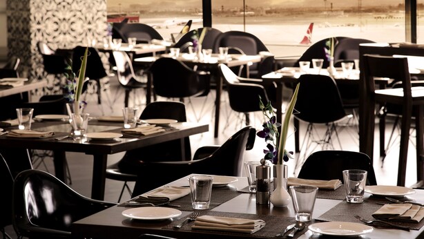 Un restaurante con mesas dispuestas para comer, vistas a la playa y un avión en la distancia.
