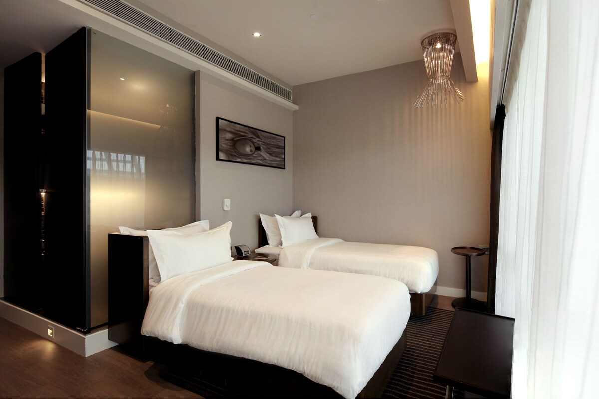 A modern hotel room with two beds, a large window with curtains, a wall-mounted artwork, and a ceiling light fixture.