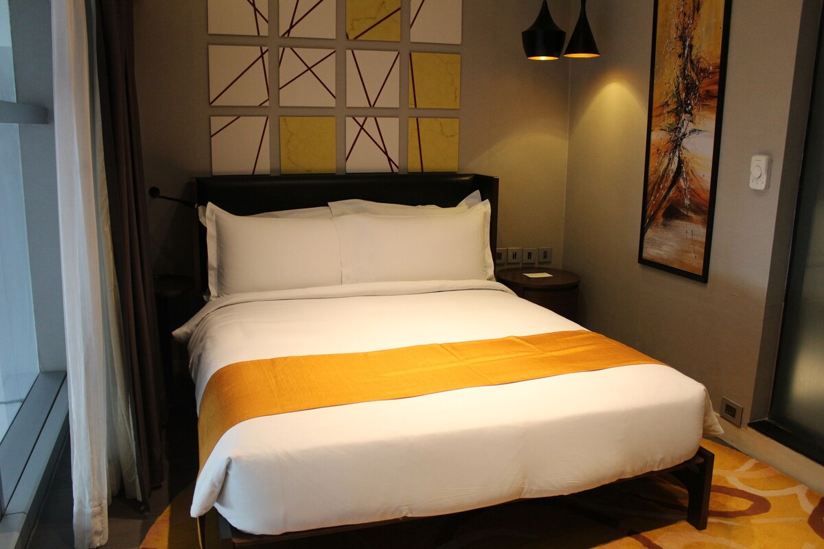 A neatly made bed with white linens and a yellow accent, a bedside table with a lamp, and a wall with geometric decor.