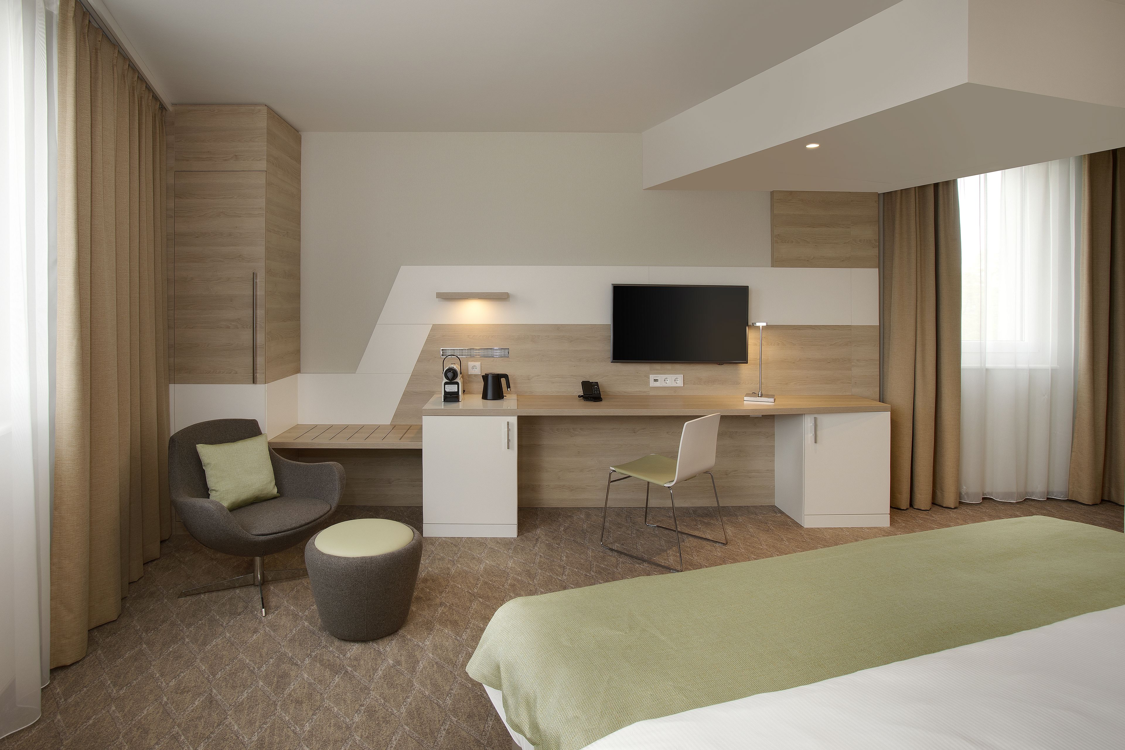 A modern hotel room with a bed, a desk with a flat-screen TV, a chair, a small cabinet, and a coffee machine.