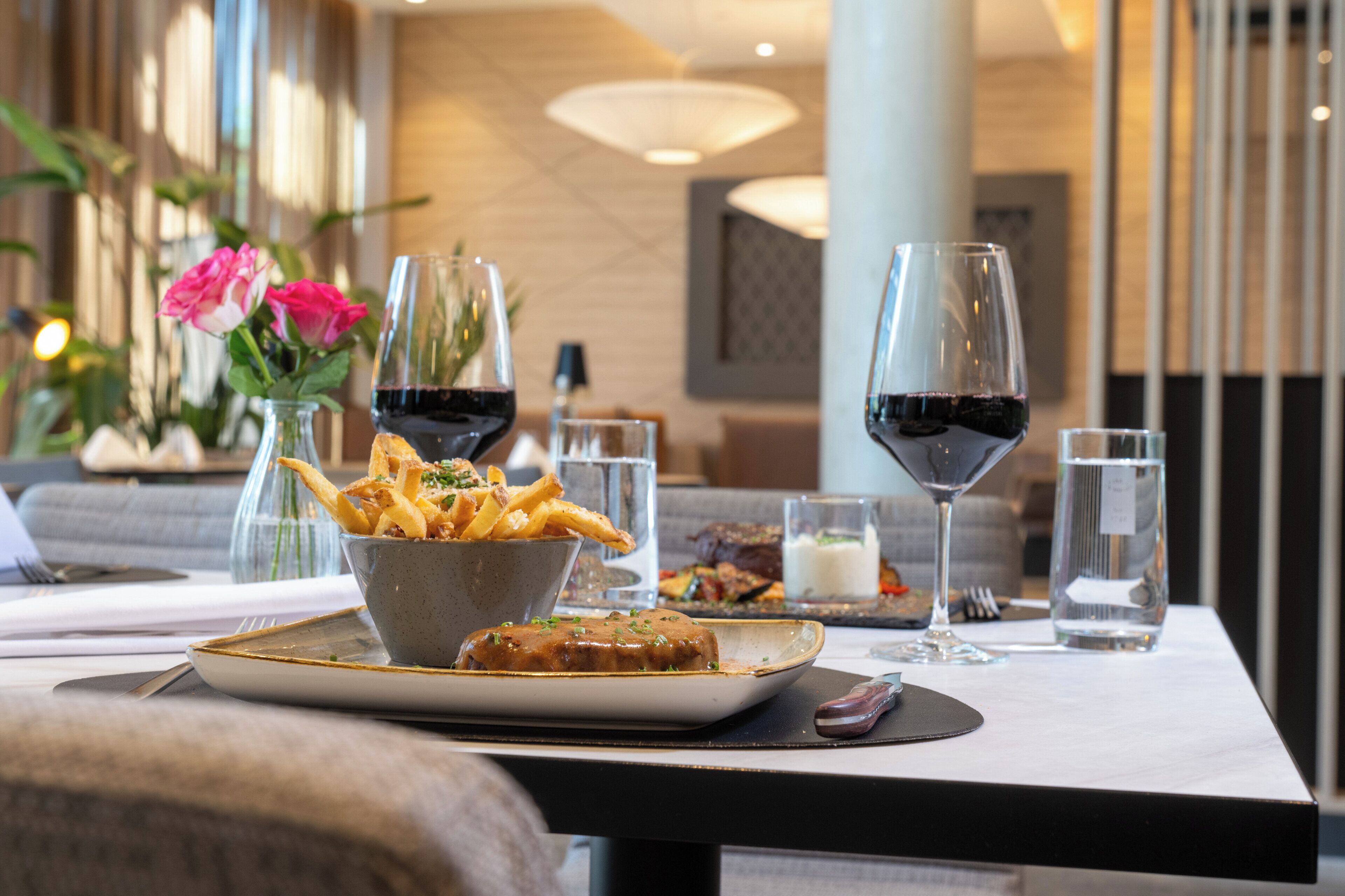Una mesa de comedor con un plato de comida, un bol de papas fritas y una copa de vino tinto.