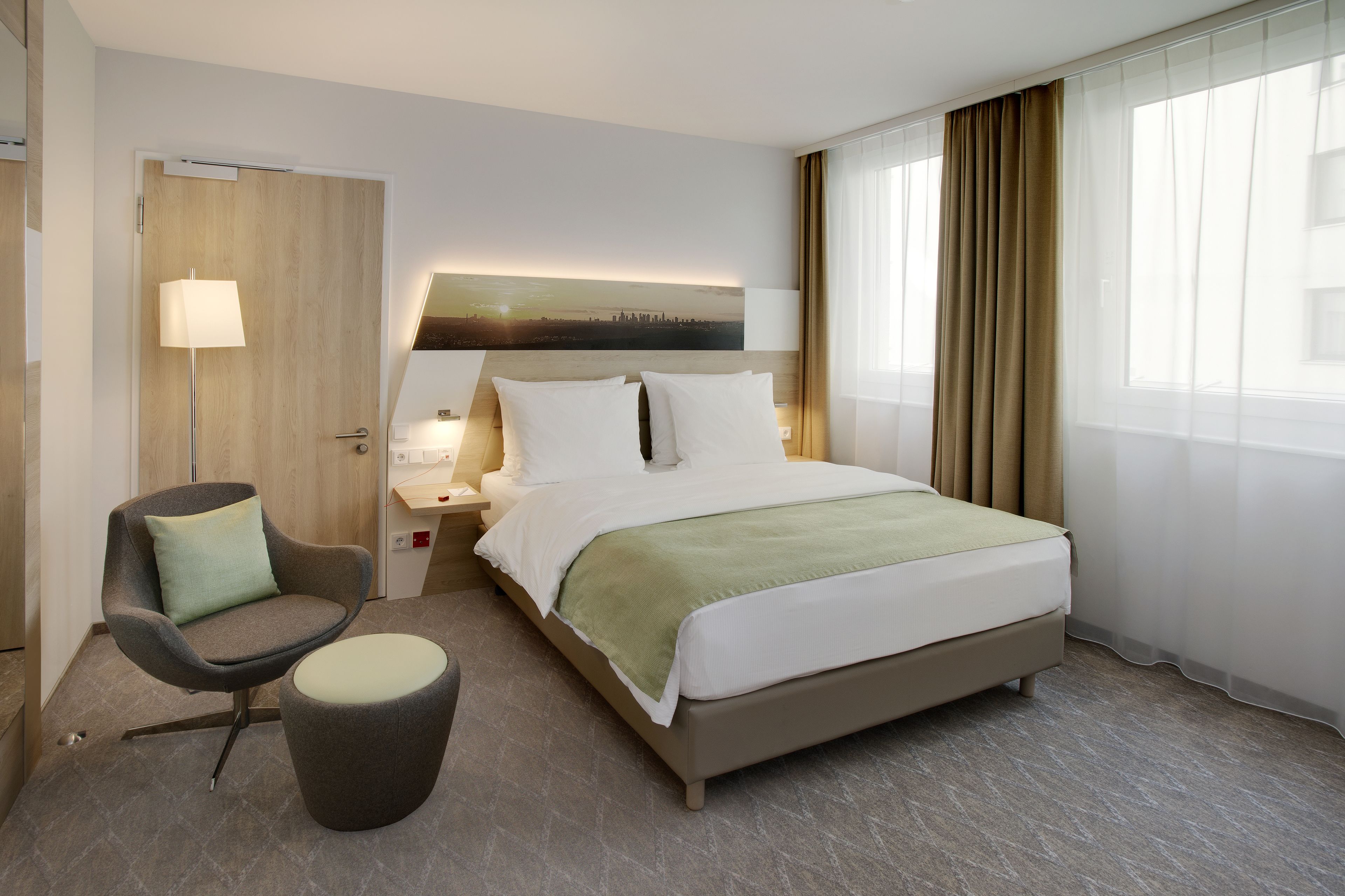 A hotel room with a bed, a chair, a nightstand, a lamp, and a view of the city skyline through sheer curtains.