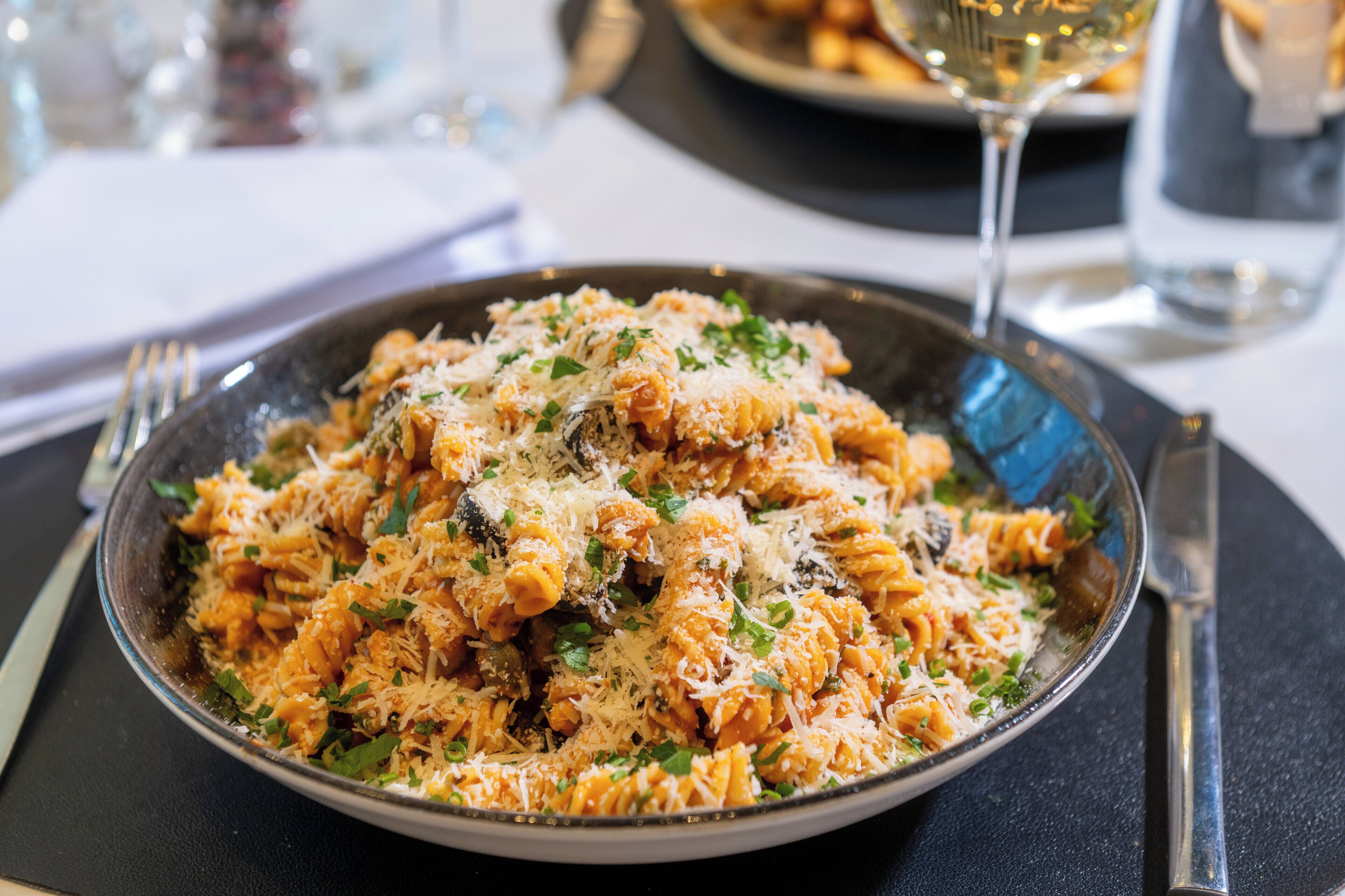 Un plato de pasta con queso y hierbas, un tenedor y un cuchillo sobre una mesa.
