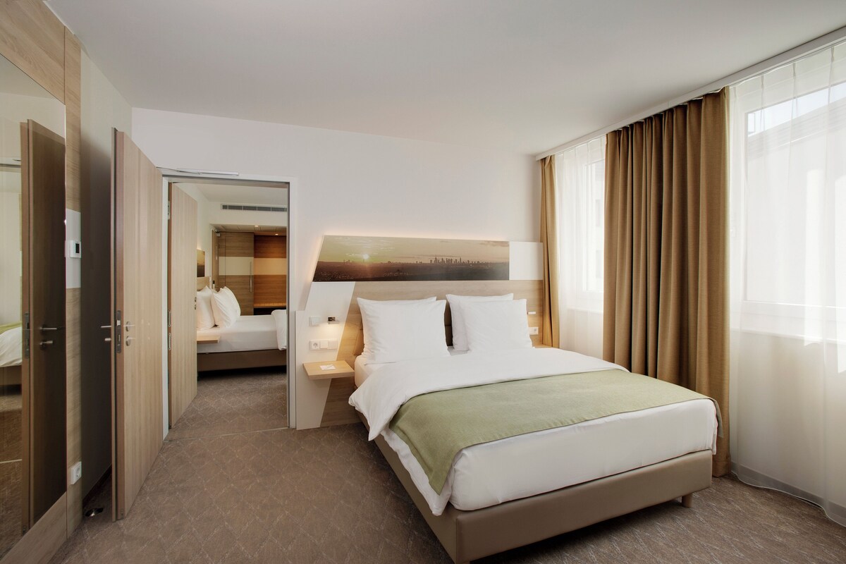 A hotel room with a bed, bedside table, and a view of a cityscape through the window.