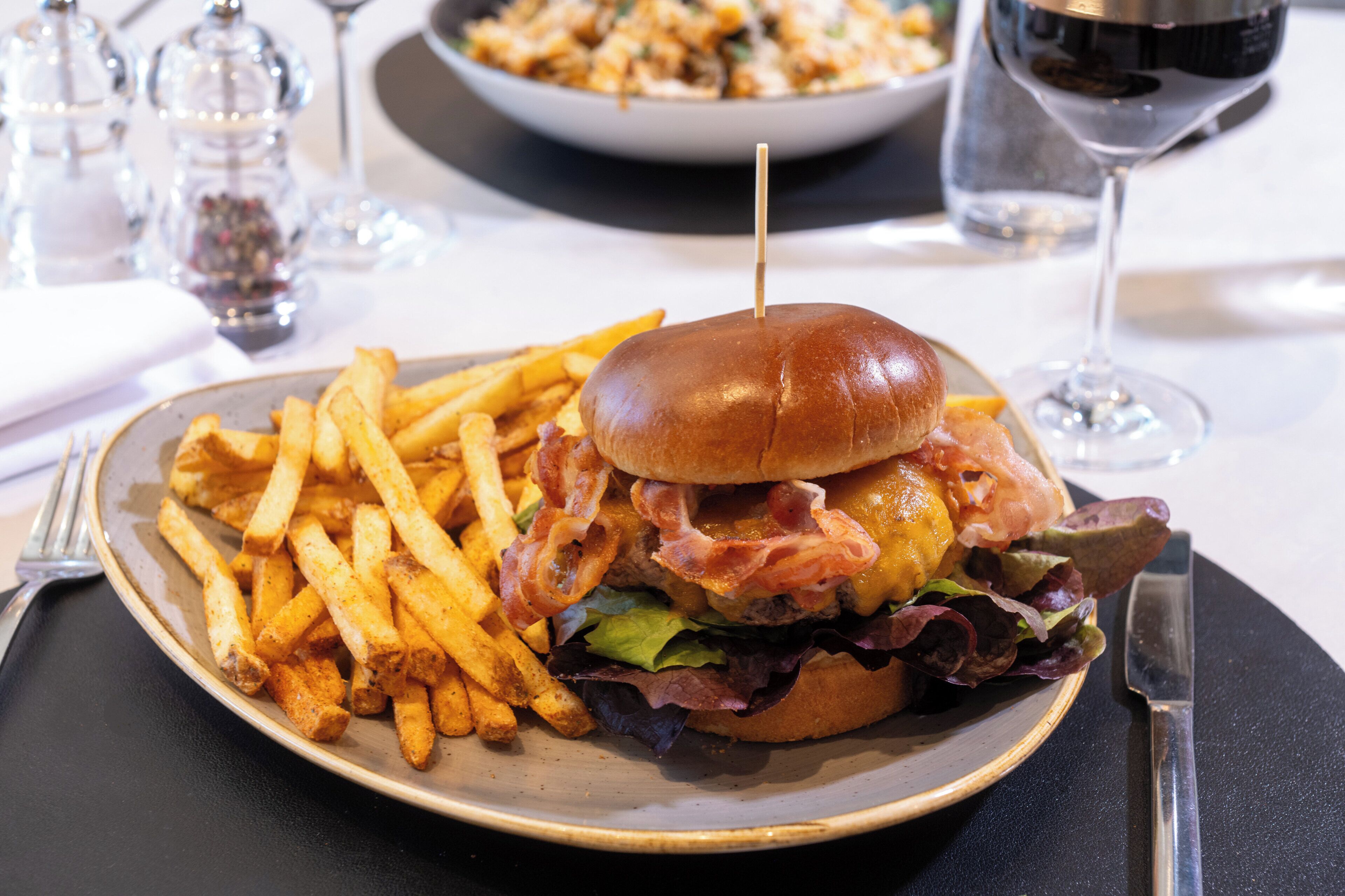 Un plato con una hamburguesa, papas fritas y una copa de vino tinto sobre una mesa.