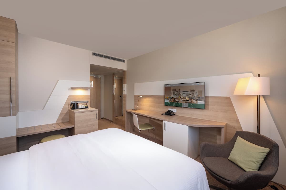 A hotel room with a bed, a desk, a chair, a lamp, and a wall-mounted TV.