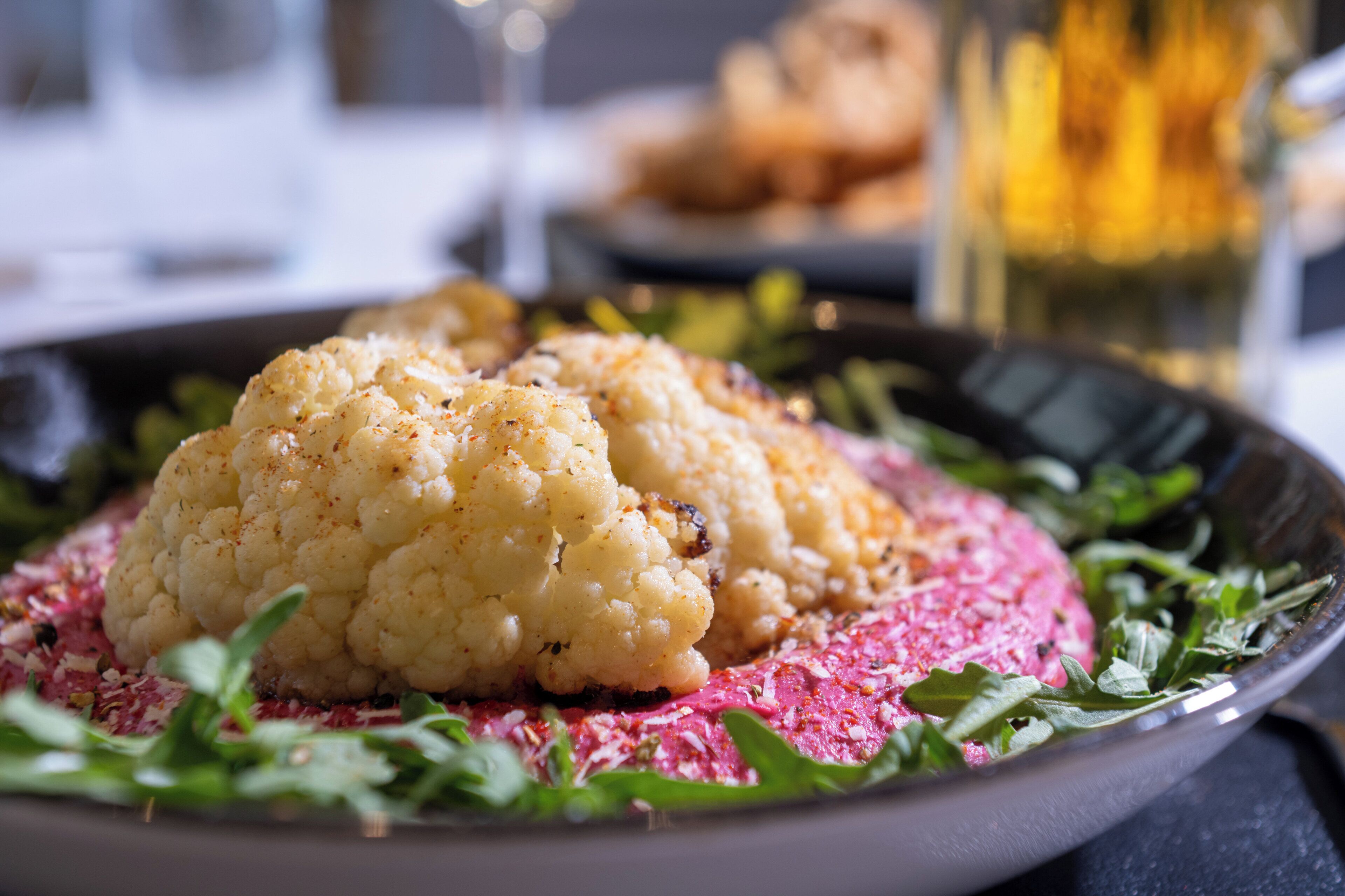 A plate with cauliflower and greens, a glass of a yellowish drink, and a blurred background with a plate of food.