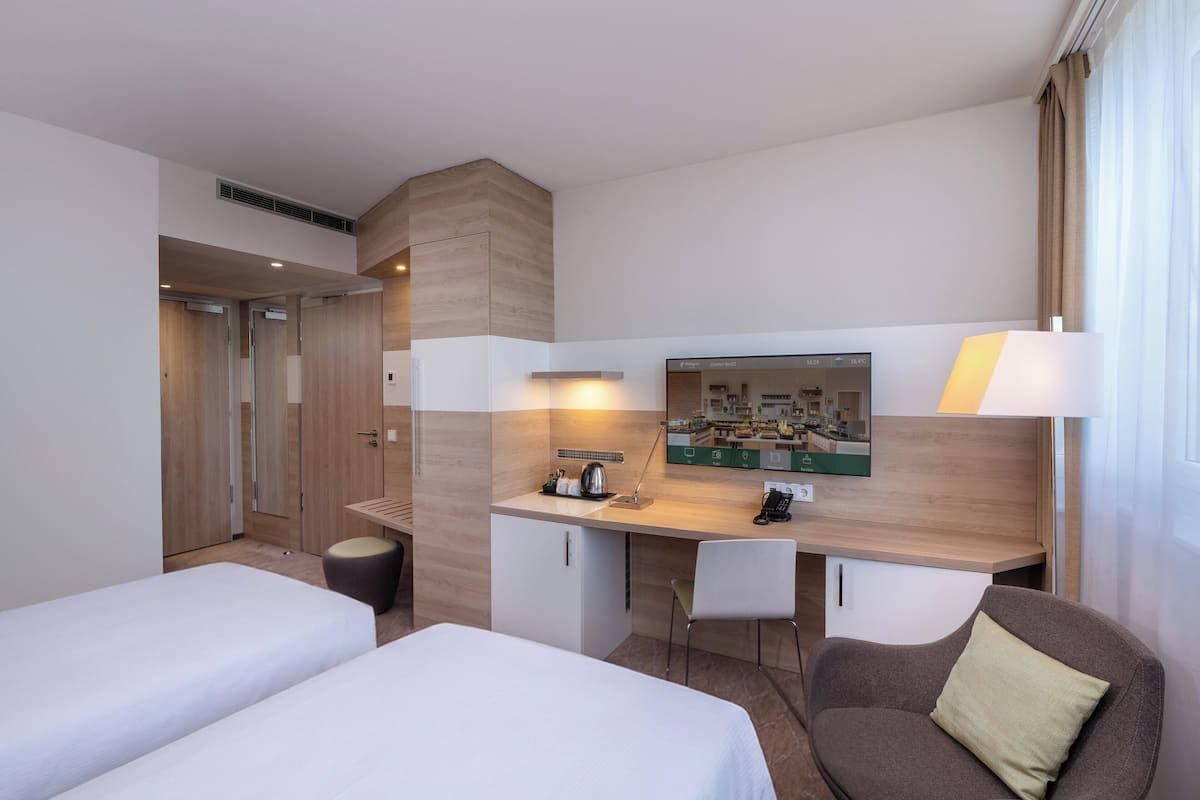 A hotel room with two beds, a desk with a chair, a small table, a mirror, and a lamp.