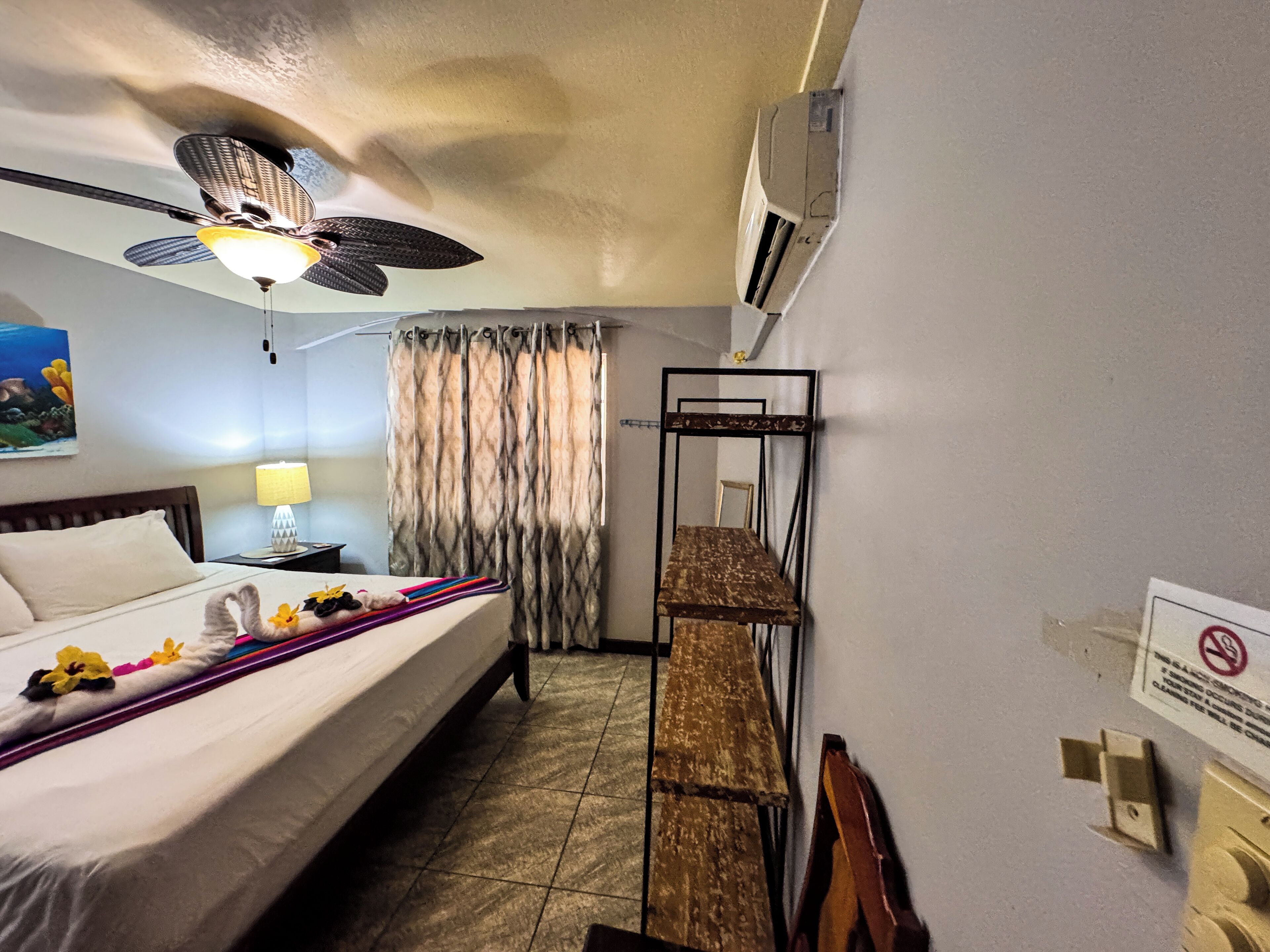 Une chambre d’hôtel équipée d’un lit, d’un bureau, d’un ventilateur de plafond, d’une lampe, d’un rideau et d’un climatiseur fixé au mur.