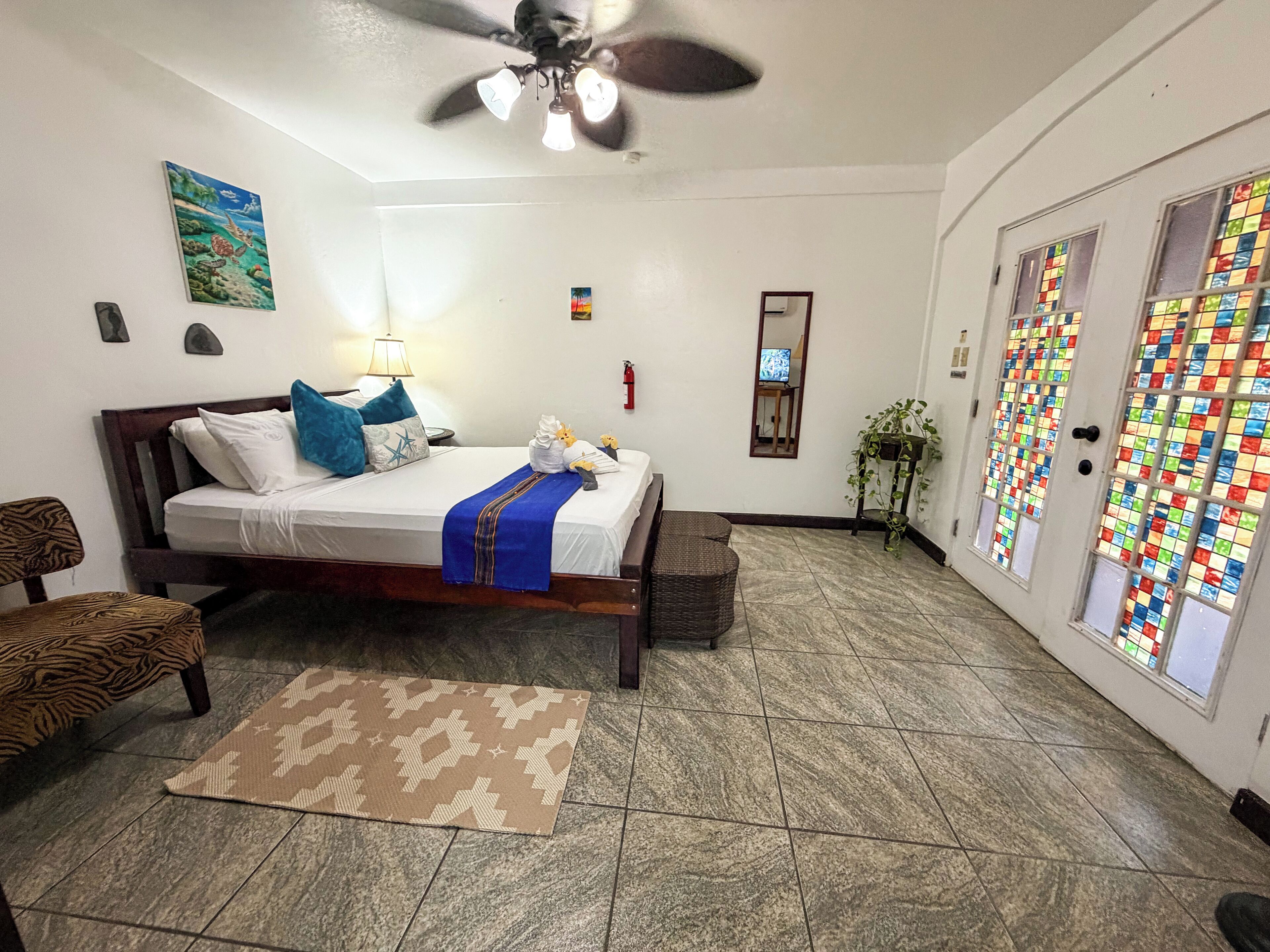 A hotel room with a bed, a ceiling fan, a TV, a window with a colorful view, and a door with a patterned glass design.