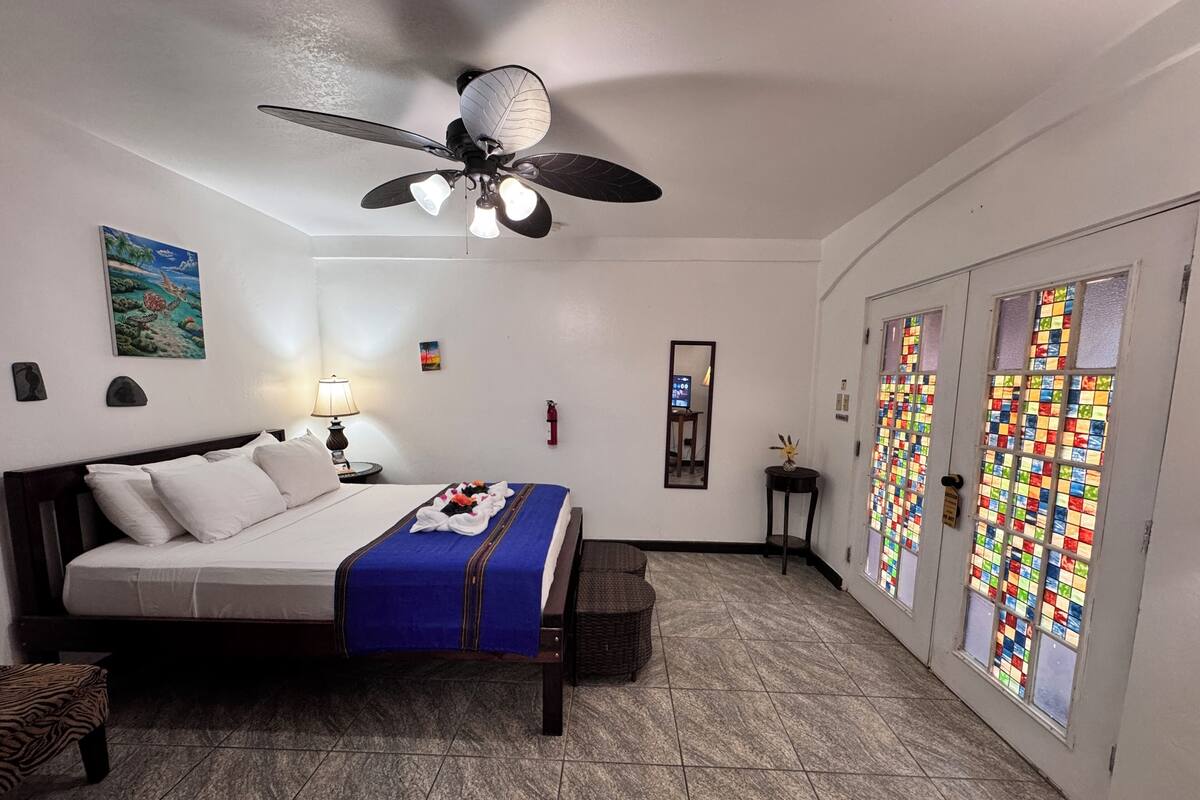 A hotel room with a bed, a ceiling fan, a nightstand, a lamp, a small table with a vase, a fire extinguisher, a mirror, and a door with colorful stained glass windows.
