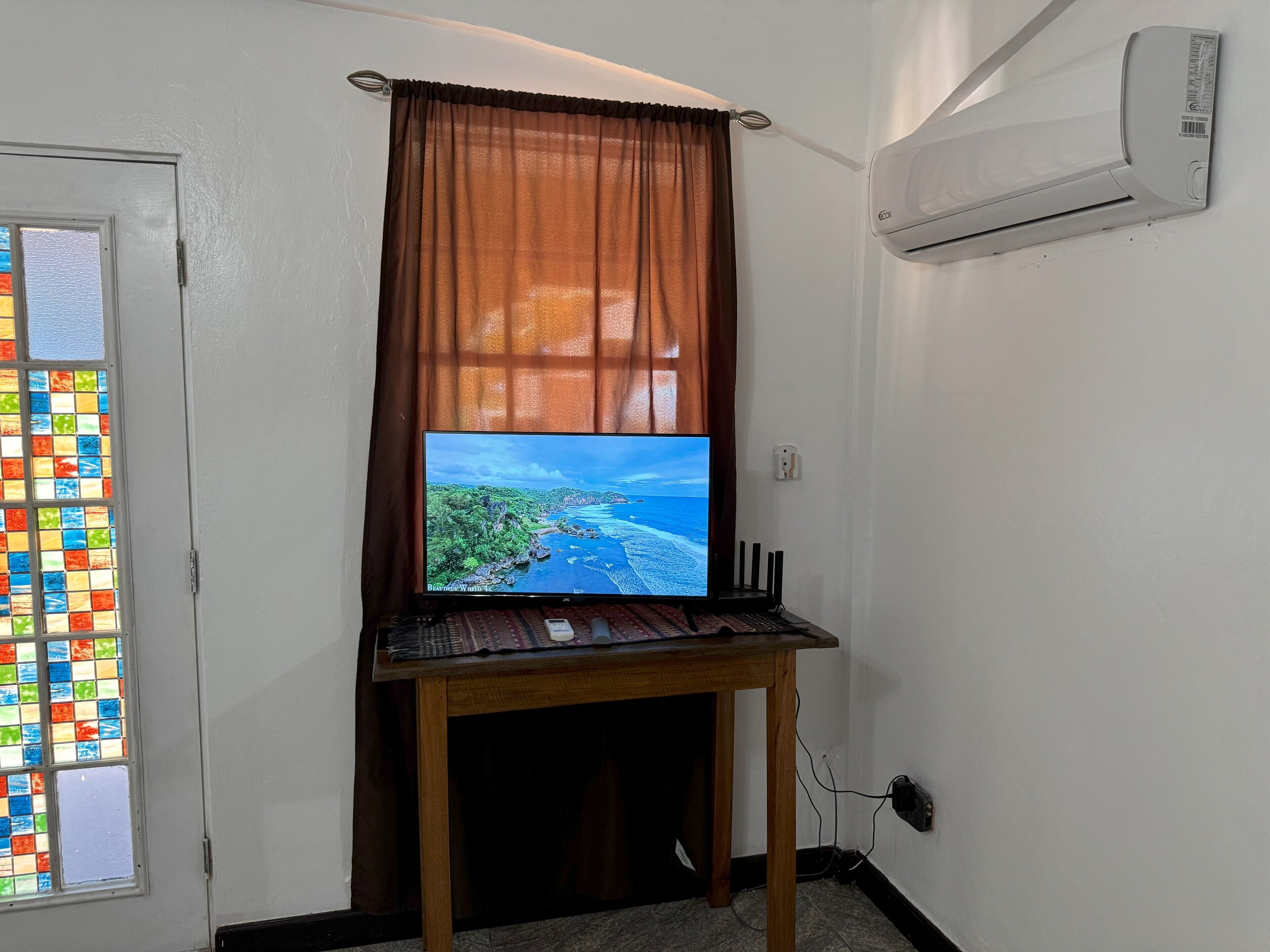 Una habitación con un mueble bajo de madera que sostiene un televisor de pantalla plana, una ventana con vidrieras de colores y un equipo de aire acondicionado en la pared.