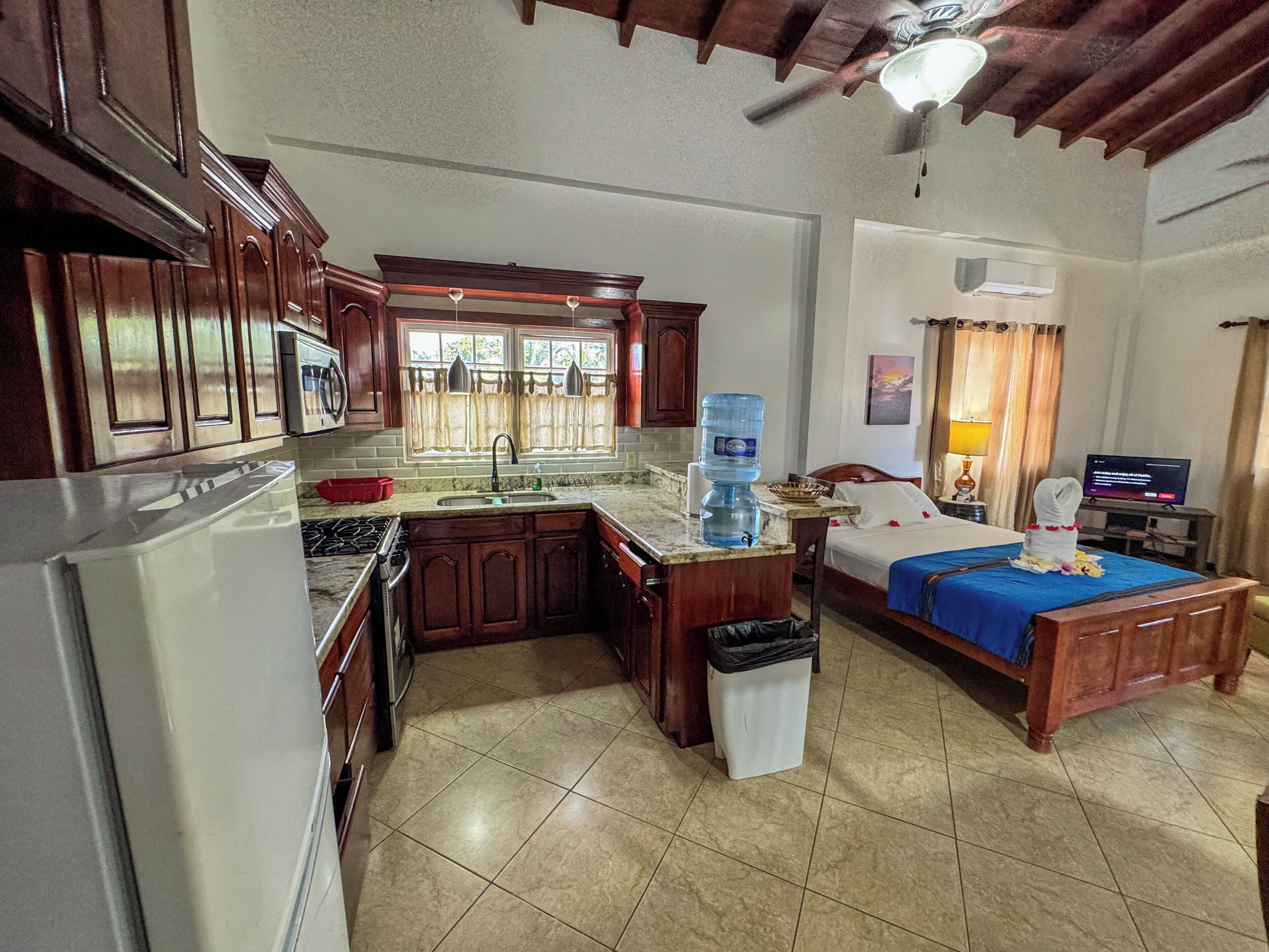 A compact hotel room with a kitchenette, a bed with a blue bedspread, a TV, and a water dispenser.