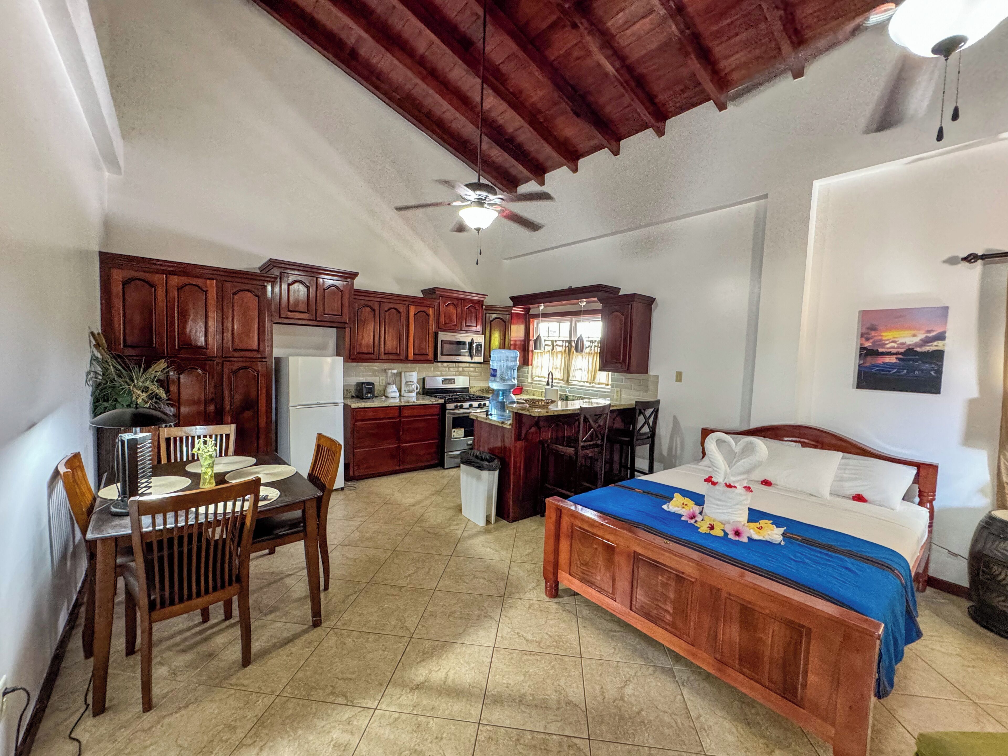 Quarto de hotel pequeno com cama de madeira, área de refeições com mesa e cadeiras, cozinha compacta e ventilador de teto.