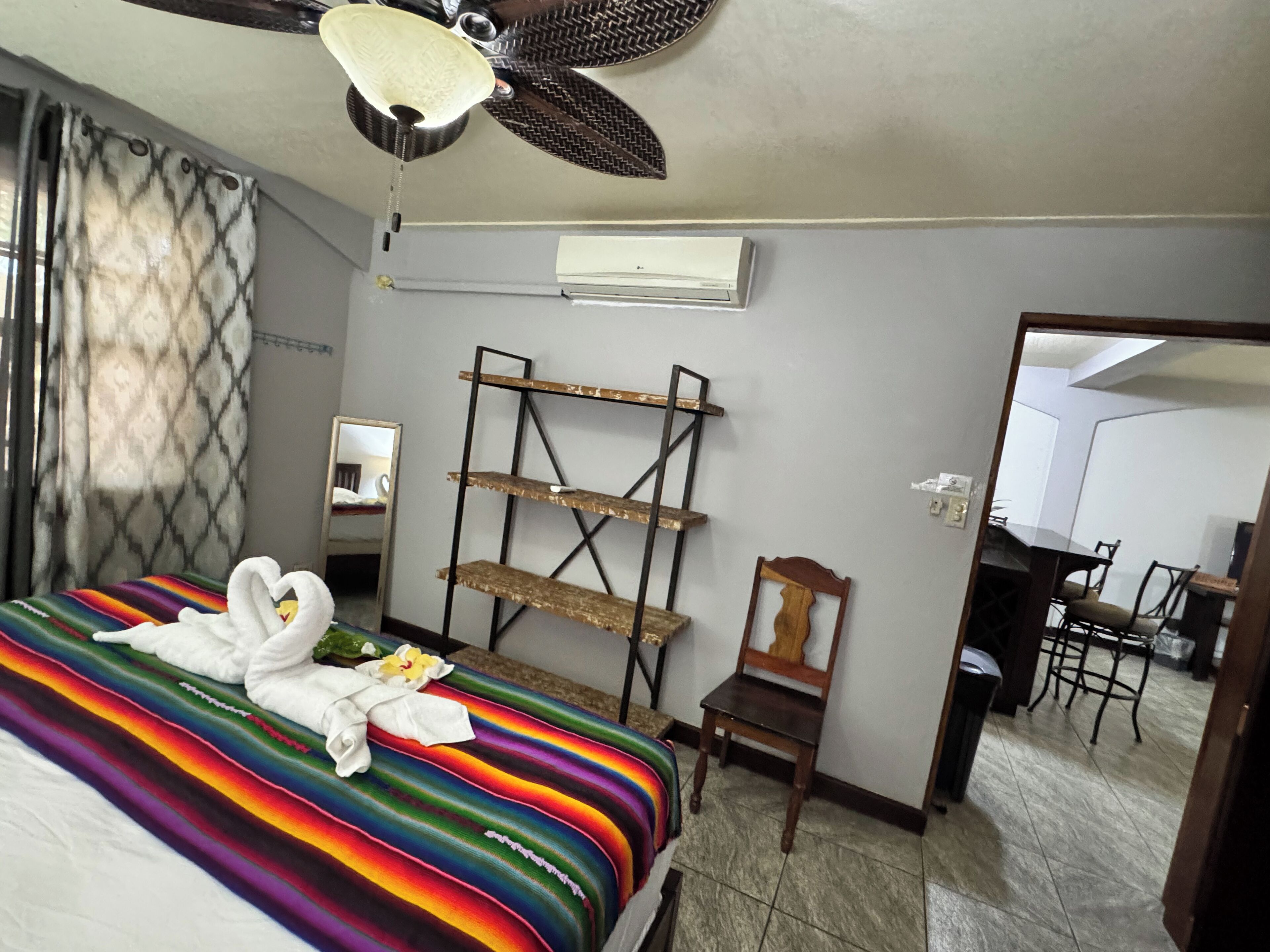 A hotel room with a bed, a ceiling fan, a mirror, a wooden chair, a desk, and a dining area.
