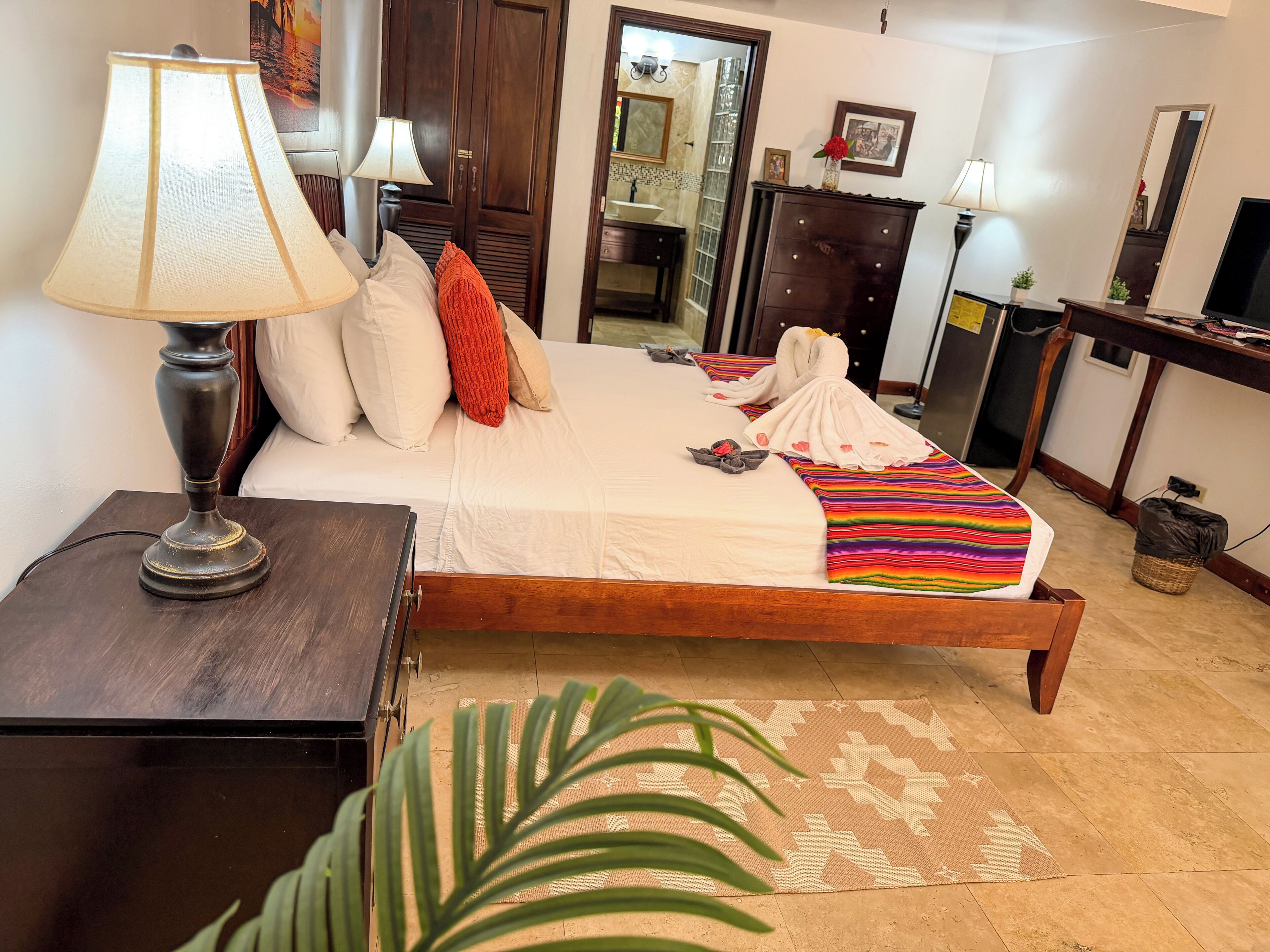 A hotel room with a bed, a nightstand, a lamp, a dresser, a mirror, and a plant.