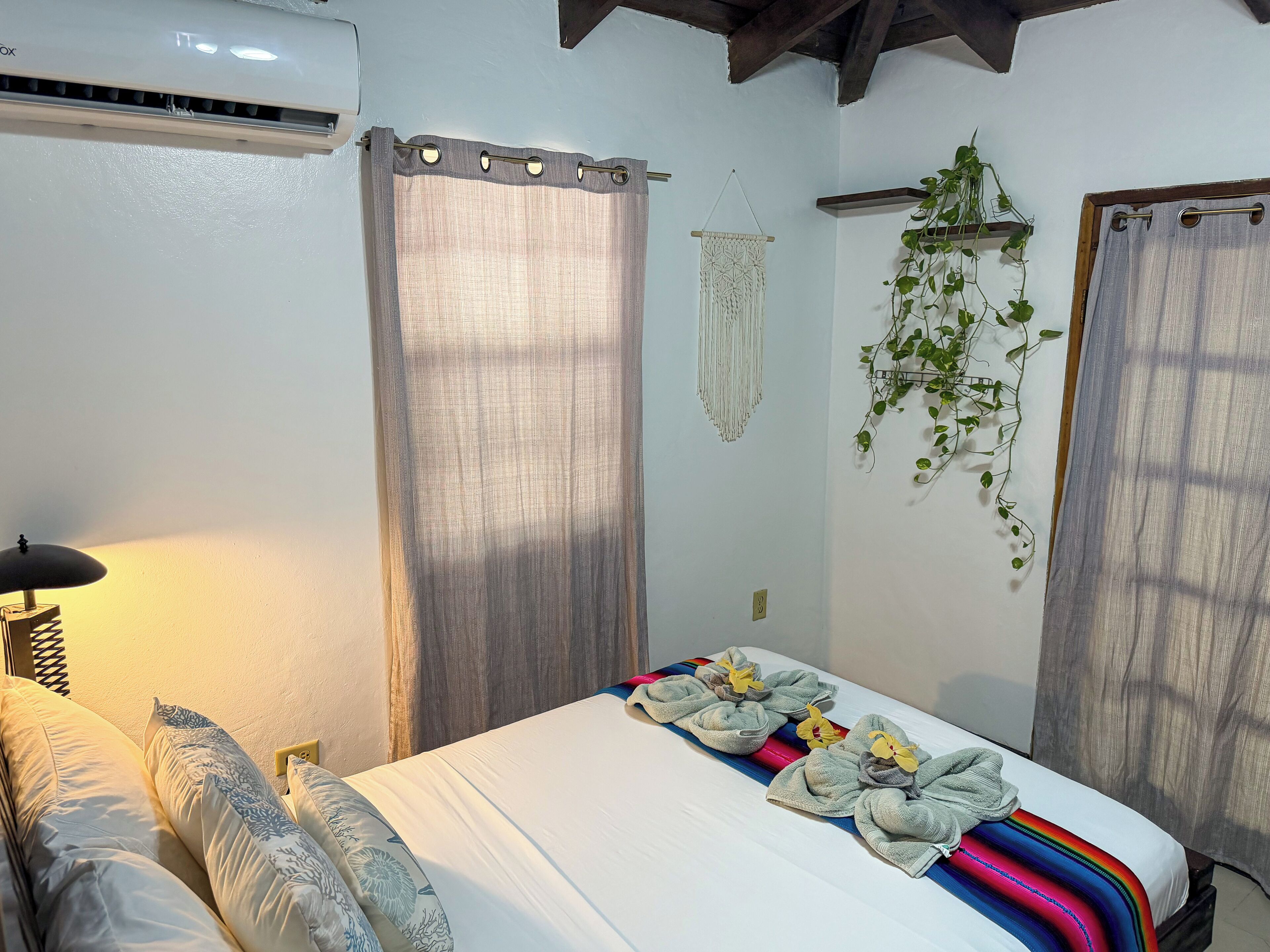 Habitación de hotel con cama, lámpara de noche, cortinas, un tapiz de macramé y una planta colgante.