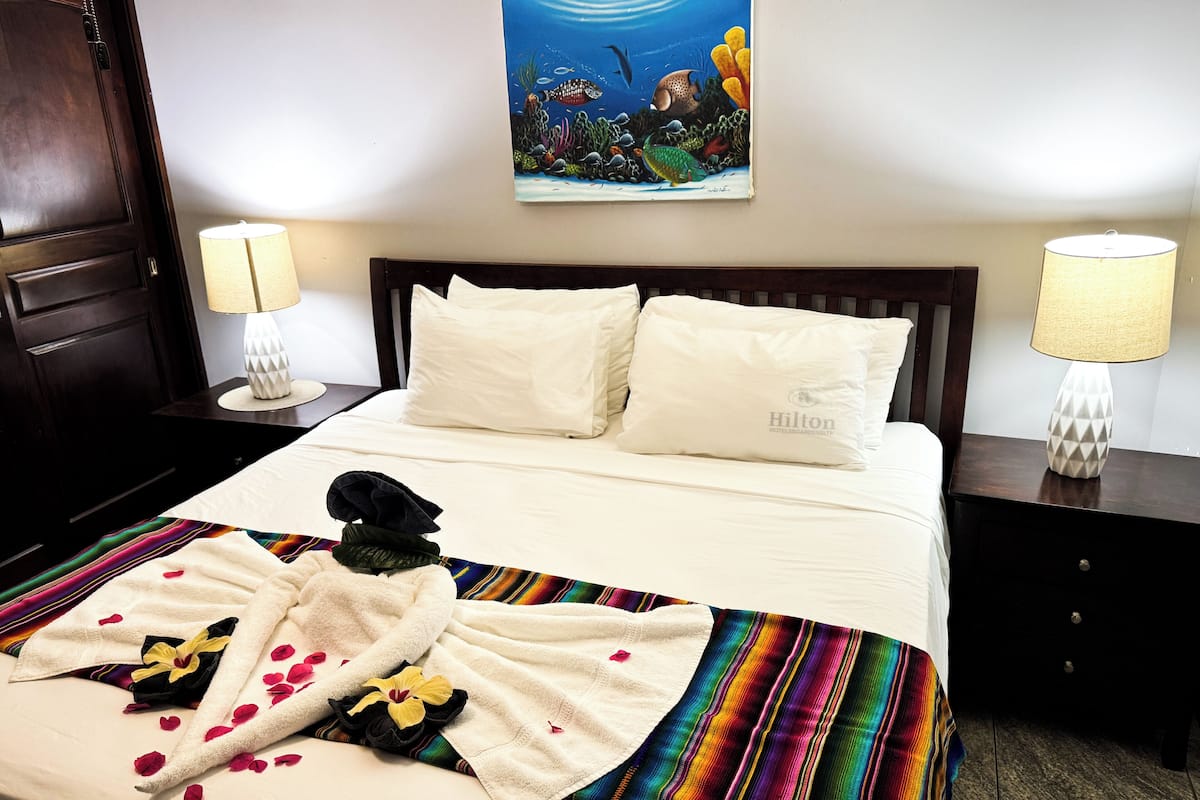 A hotel room with a bed, white pillows, a colorful blanket, a robe, and a painting of an underwater scene.