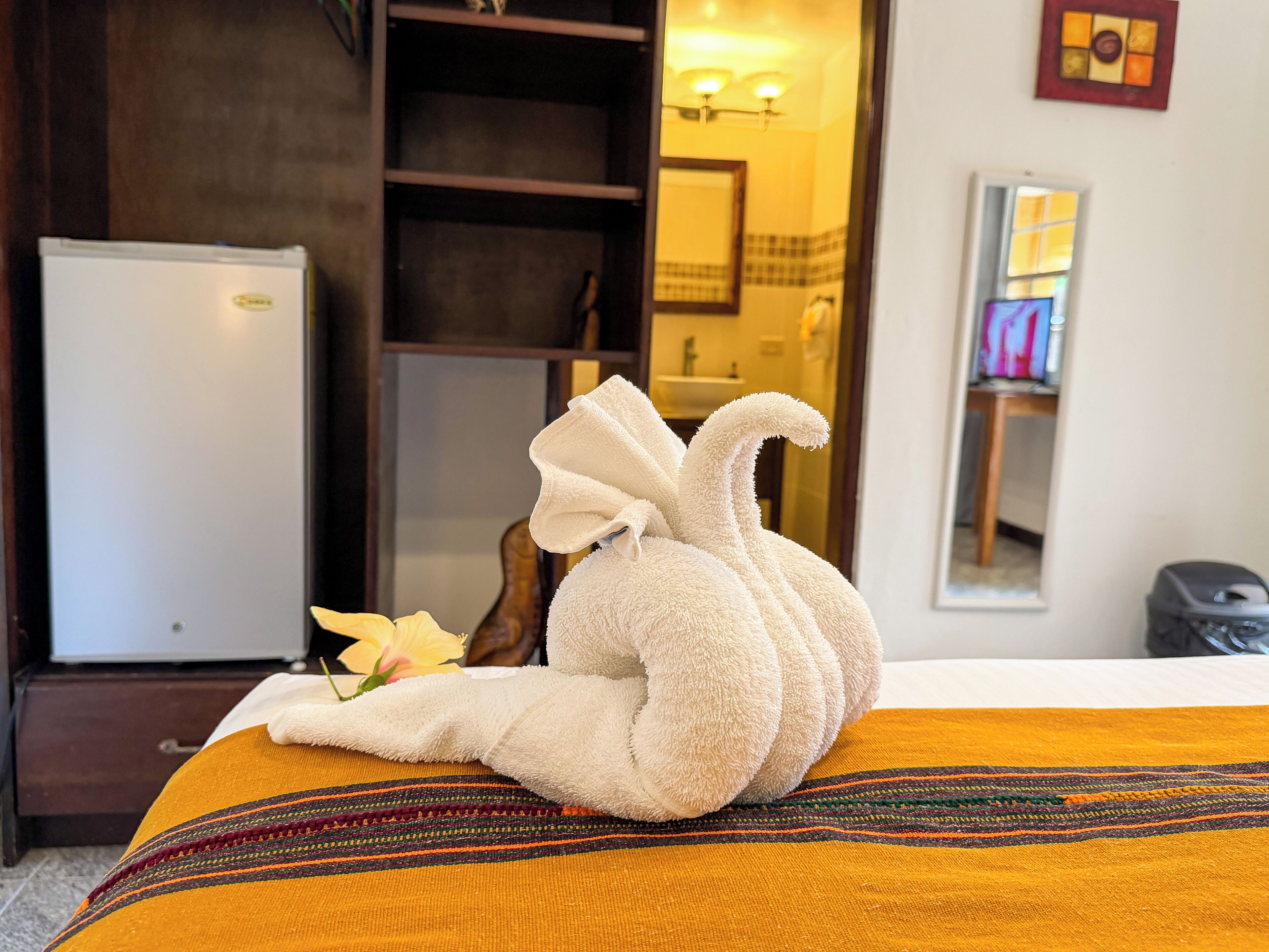 A bed with a towel shaped like a swan, a yellow-striped blanket, a white refrigerator, and a mirror reflecting a bathroom.