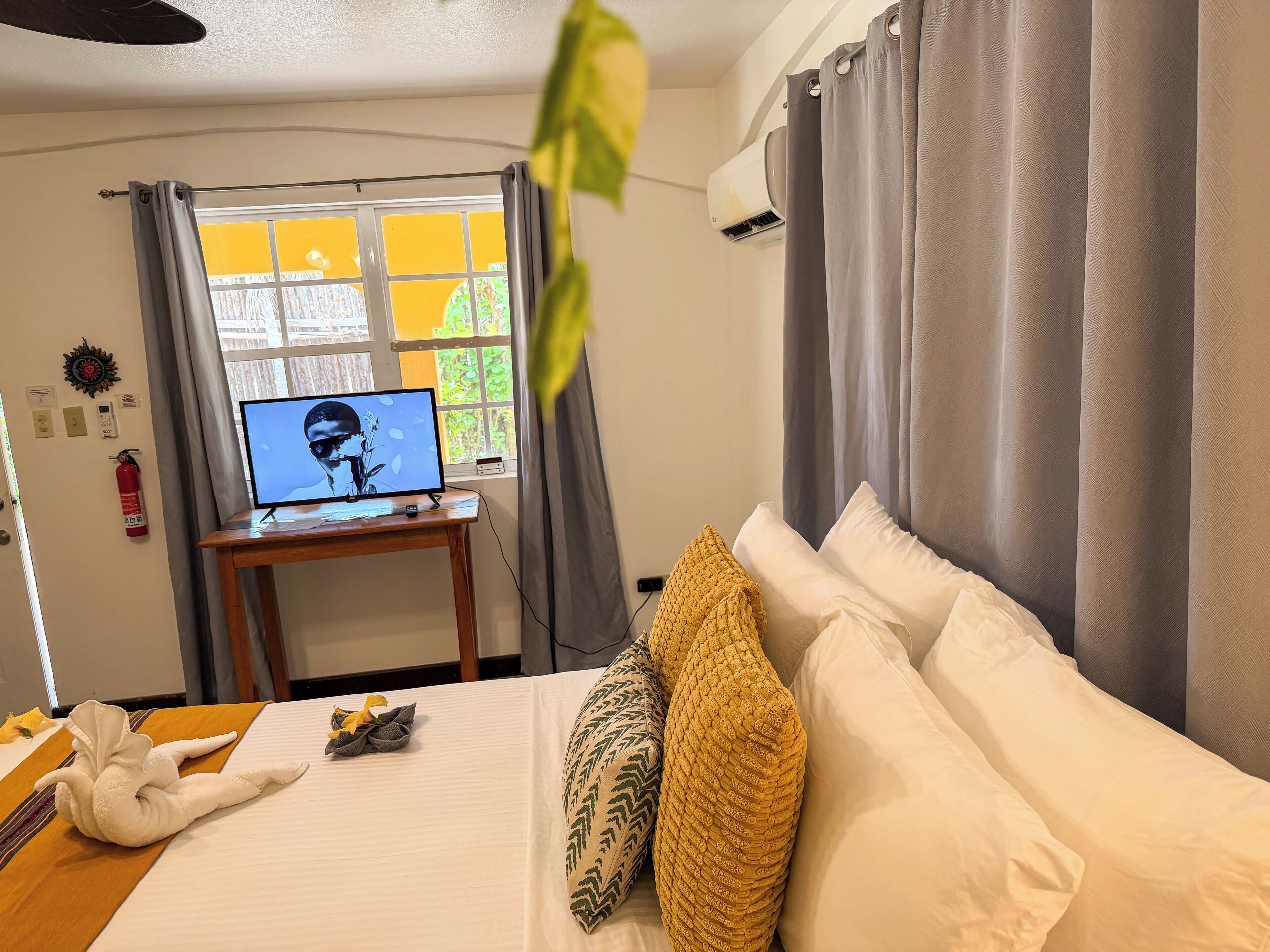 Habitación de hotel con cama, escritorio con computadora, ventana con cortinas y aire acondicionado instalado en la pared.