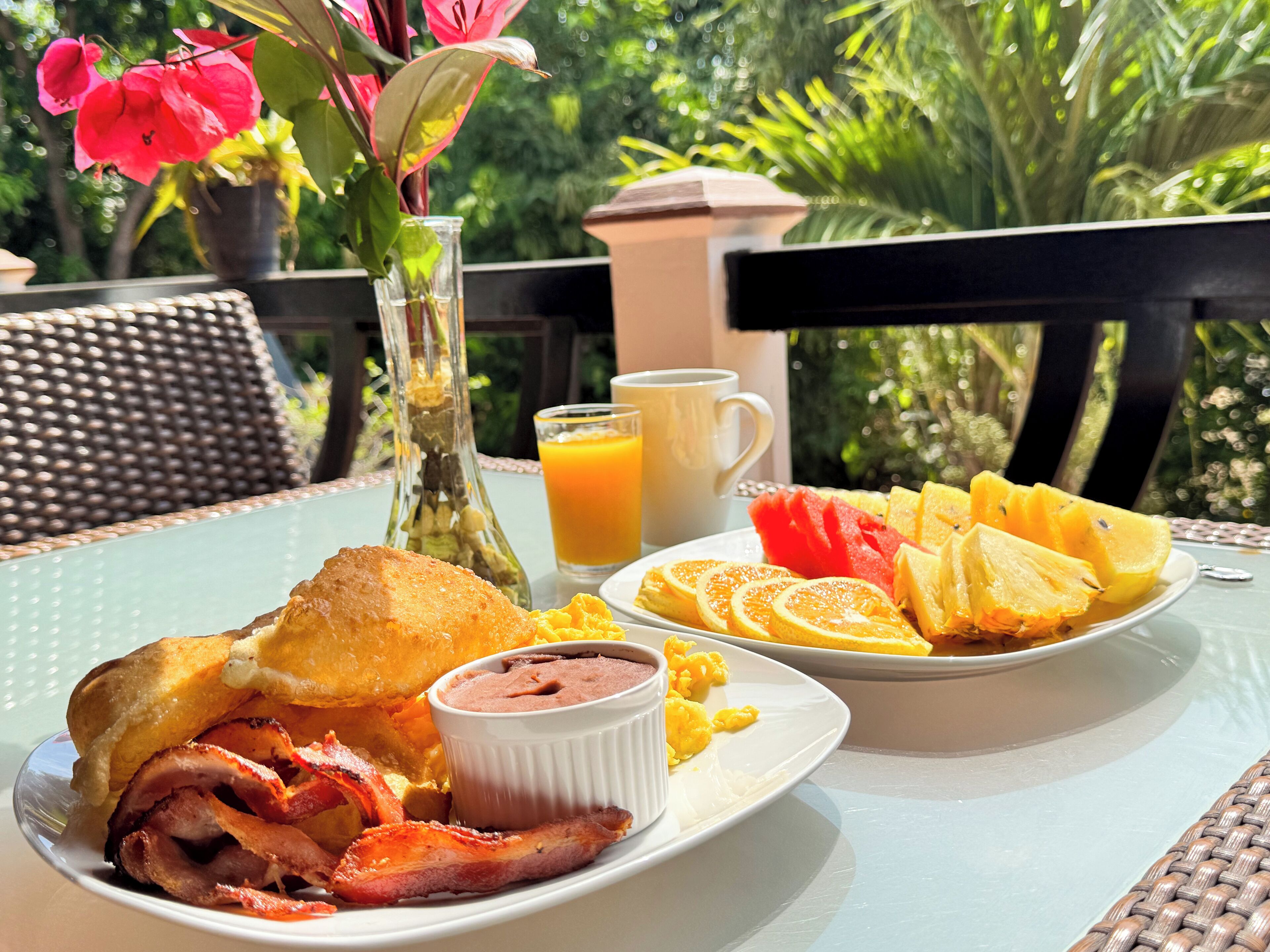 A breakfast table with a plate of eggs, bacon, toast, and fruit, a glass of orange juice, and a vase with flowers.