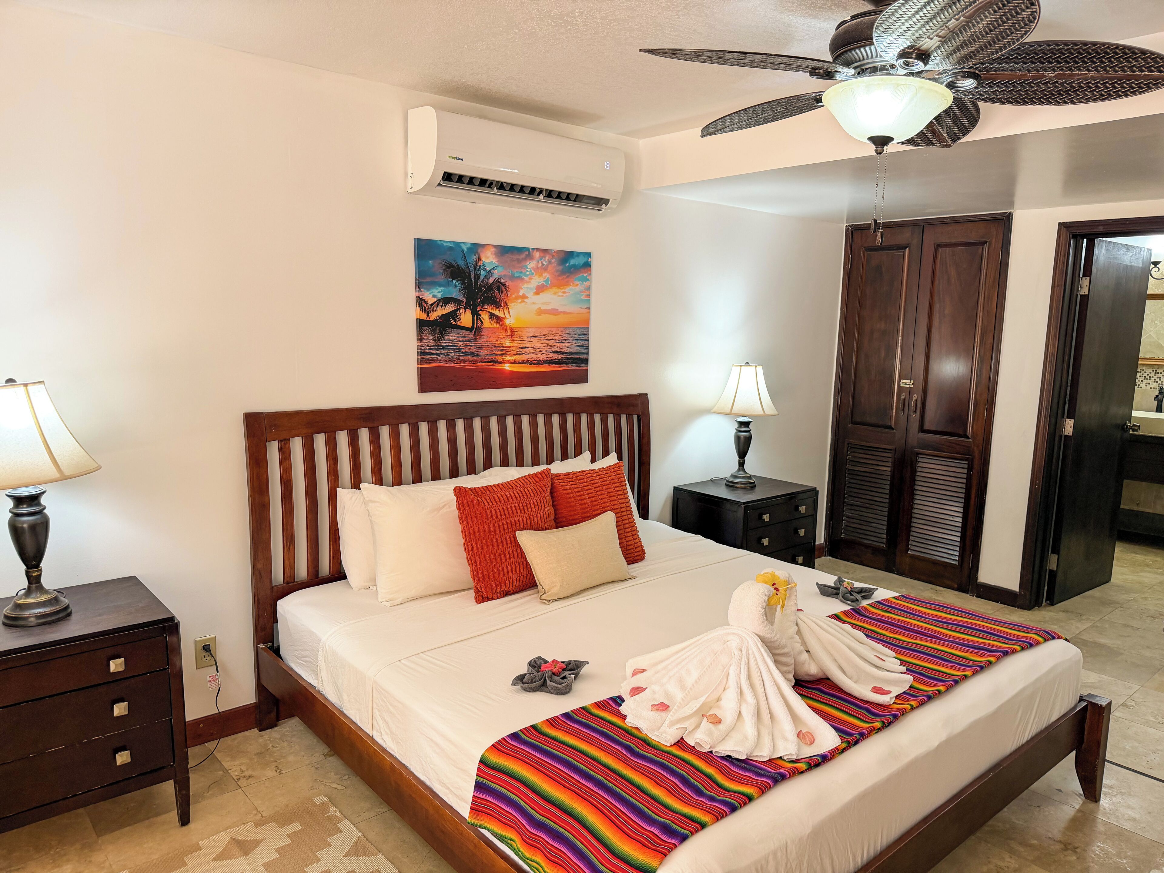 Habitación de hotel con cama, mesita de noche, lámpara, ventilador de techo, unidad de aire acondicionado y un cuadro en la pared.