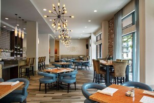 Un restaurante moderno con mesas de madera y sillas azules, una zona de bar con botellas, y una gran lámpara de araña.