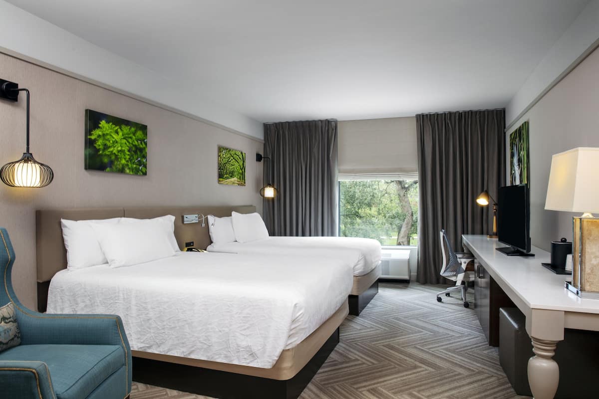 A hotel room with two beds, a desk, a chair, a TV, and a window with curtains.