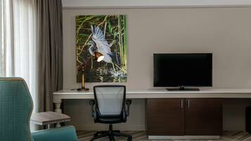 A hotel room with a desk, a television, a chair, and a painting of a bird.