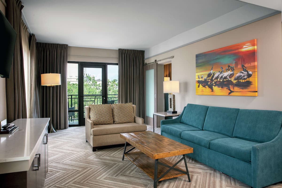 A hotel room with a sofa, a coffee table, a TV, a painting, and a view of greenery through the window.