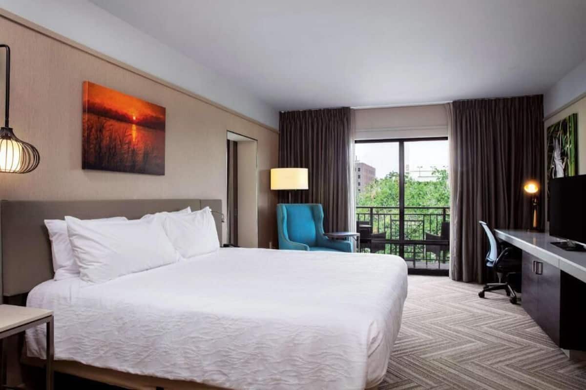 A hotel room with a bed, a desk, a chair, a TV, and a balcony with curtains.