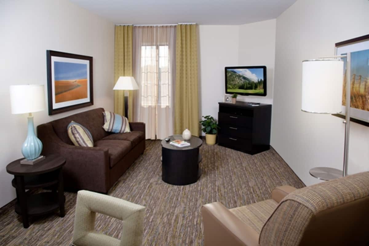 A hotel room with a brown sofa, a small round table, a TV, a lamp, and a framed picture on the wall.