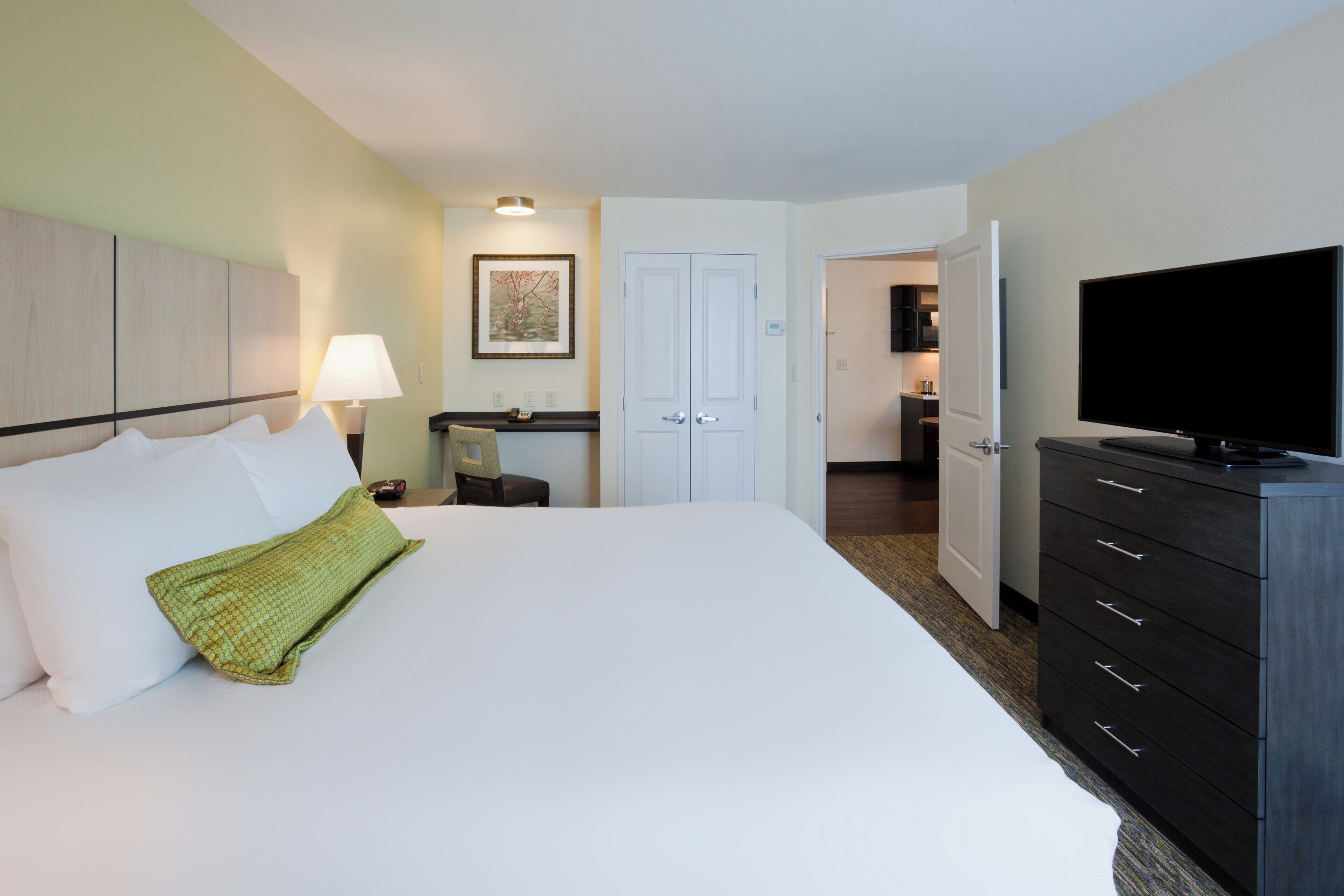 Ein Hotelzimmer mit Bett, Fernseher, Schminktisch, Schreibtisch und einer Tür zu einer Küchenzeile.