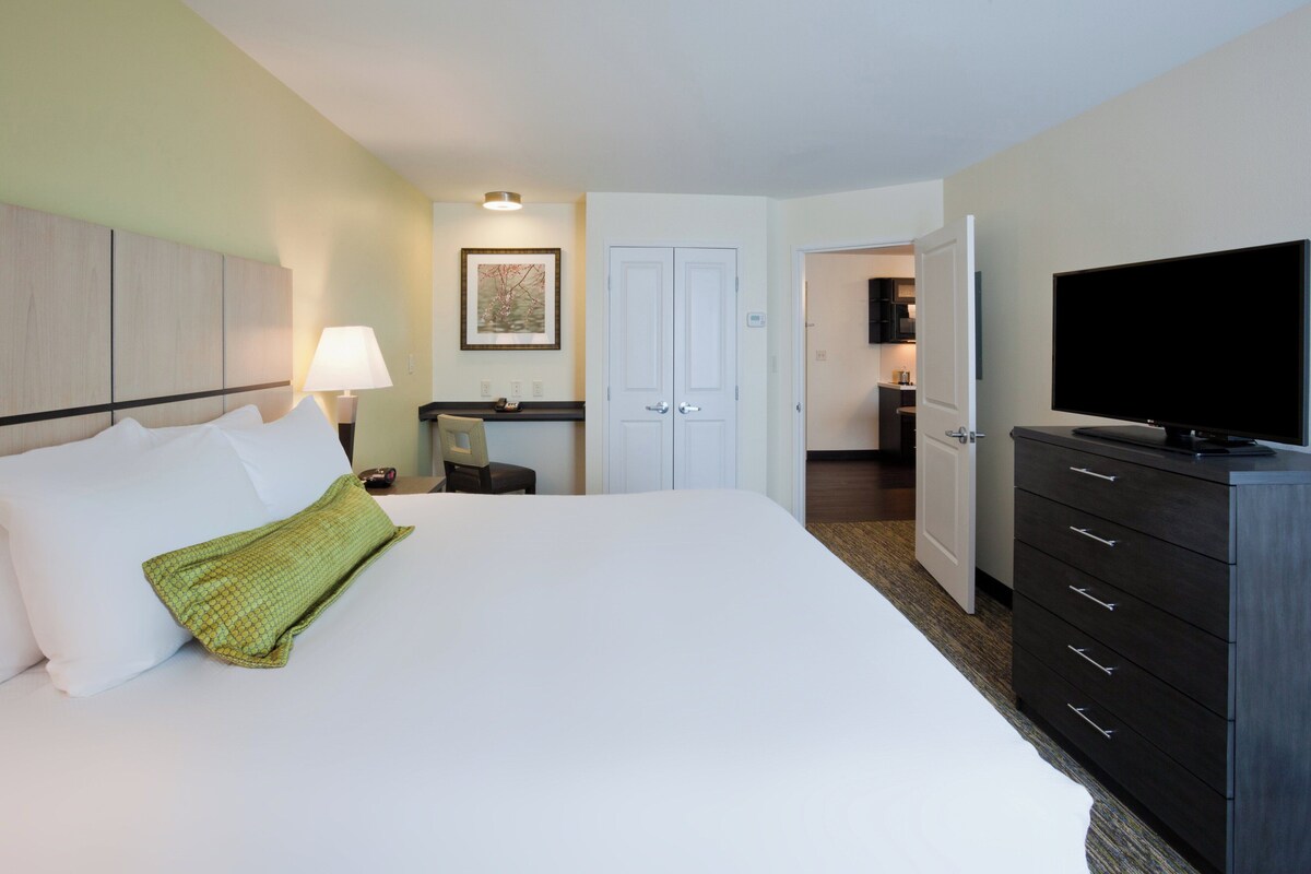 Habitación de hotel con cama, televisión, cómoda, escritorio y puerta que da a una kitchenette.