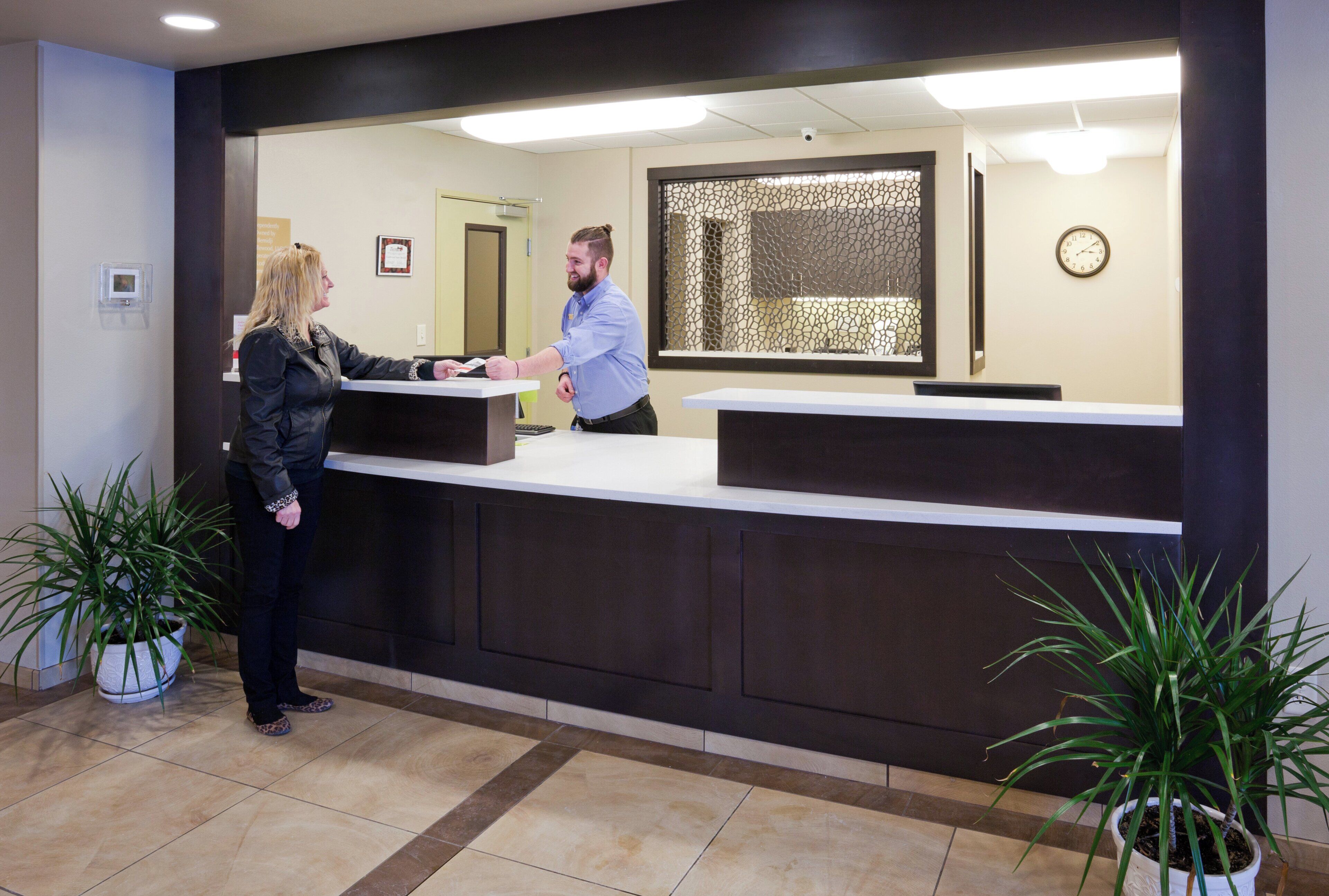 A reception desk with a person standing behind it, a potted plant to the left, and a clock on the wall.