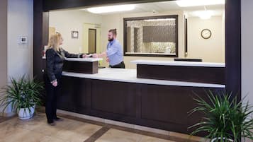 A reception desk with a person standing behind it, a potted plant to the left, and a clock on the wall.
