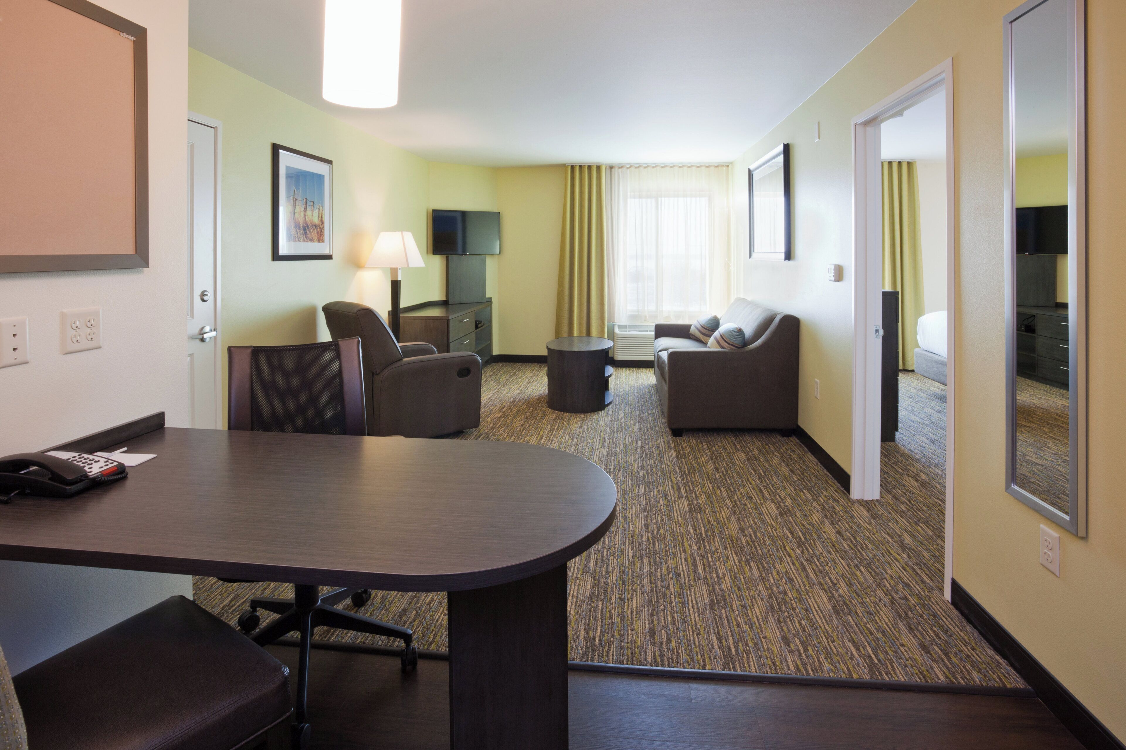 A hotel room with a conference table, two armchairs, a sofa, a TV, and a window with curtains.