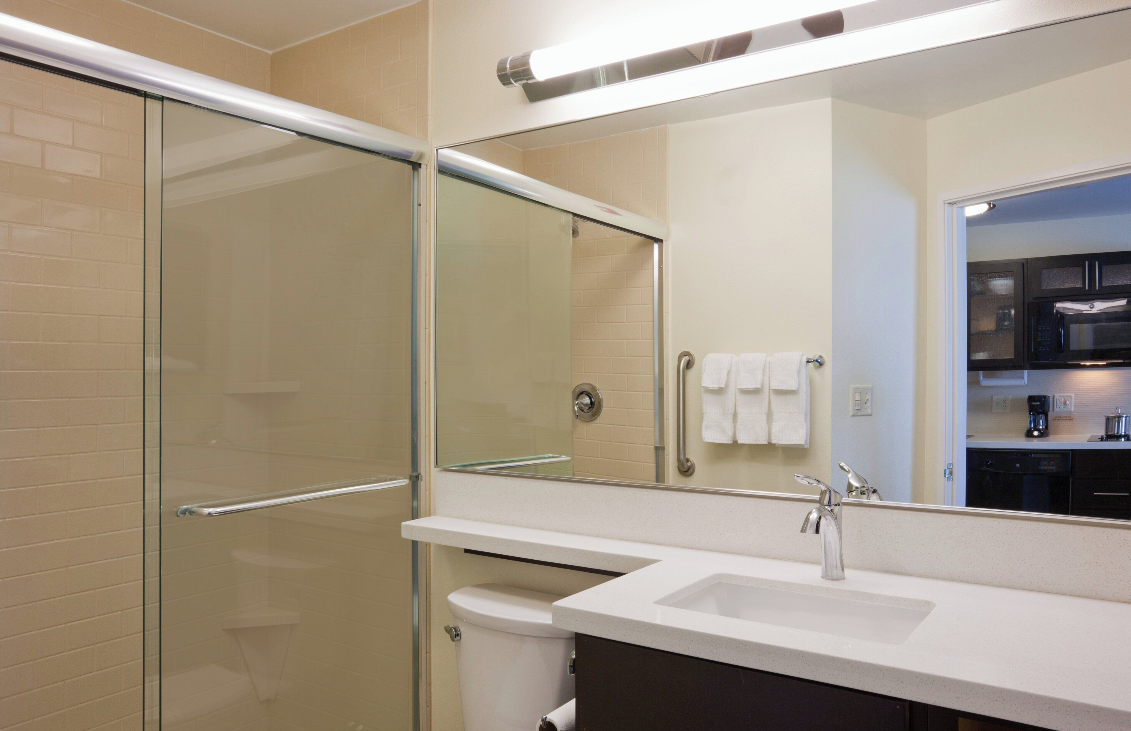 Un baño moderno con cabina de ducha de vidrio, un lavabo blanco y un espejo grande que refleja una zona de cocina.