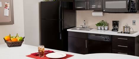 A modern kitchen with dark cabinets, a black refrigerator, a microwave, and a sink. There is a fruit basket and a glass of iced tea on the counter.