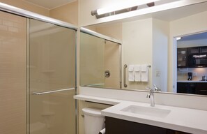 Un baño moderno con ducha envuelta en vidrio, un lavamanos blanco y un espejo que refleja una zona de cocina.