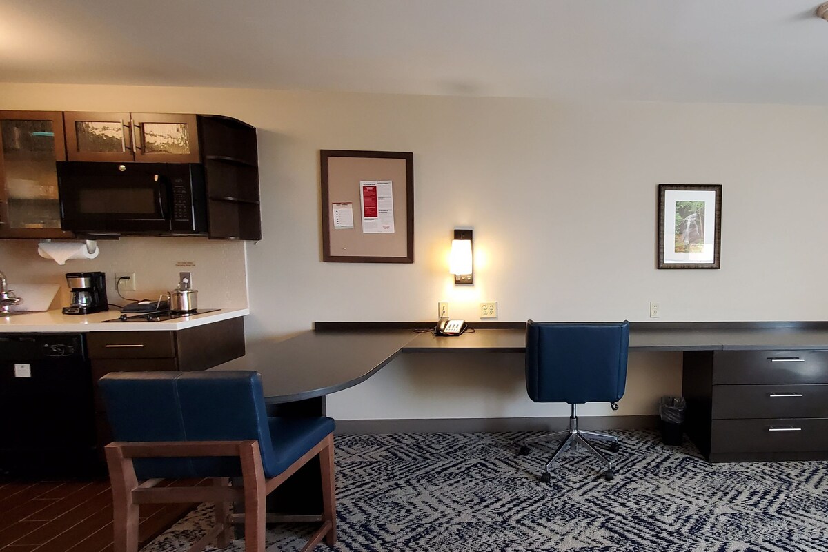 A compact hotel room with a kitchenette, a desk with two chairs, a flat-screen TV mounted on the wall, and a patterned carpet.