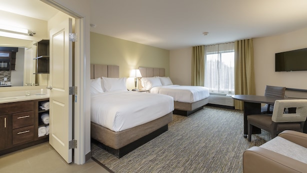 A hotel room with two beds, a desk, a chair, a TV, and a bathroom with a mirror and sink.