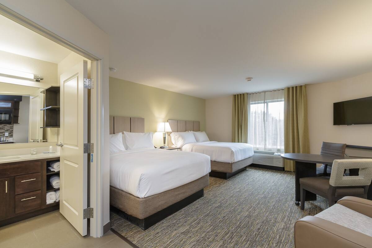 A hotel room with two beds, a desk, a chair, a TV, and a bathroom with a mirror and sink.